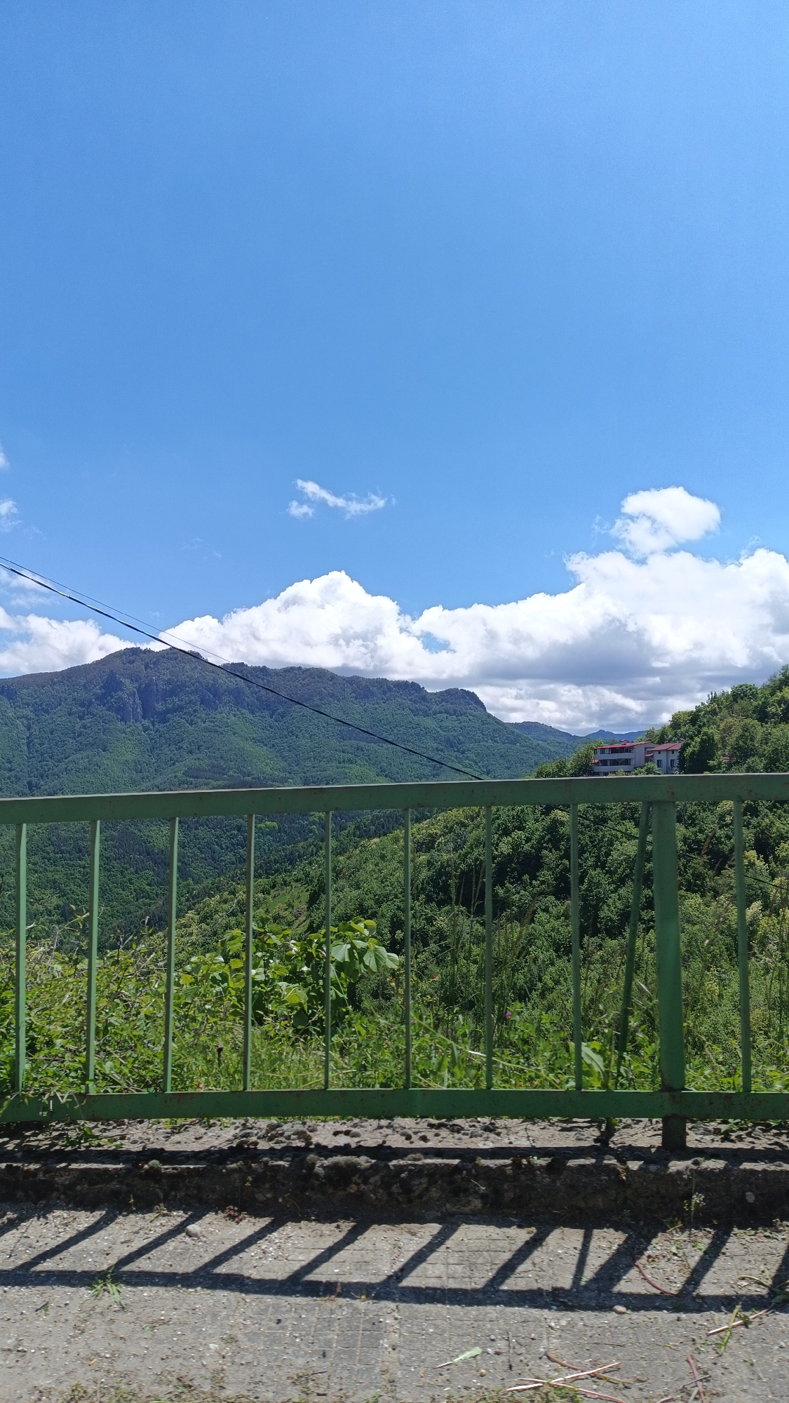 The image depicts a view from a slightly elevated position in Oreshets, Bulgaria. A green metal fence runs horizontally across the middle of the frame, casting shadows on a gray concrete surface in the foreground. Behind the fence, lush green vegetation covers a hillside that slopes downward. In the distance, a mountain range is visible, with the peak of one mountain partially obscured by clouds. The sky is blue with scattered white clouds. A building is seen on the top right side, behind the vegetation.