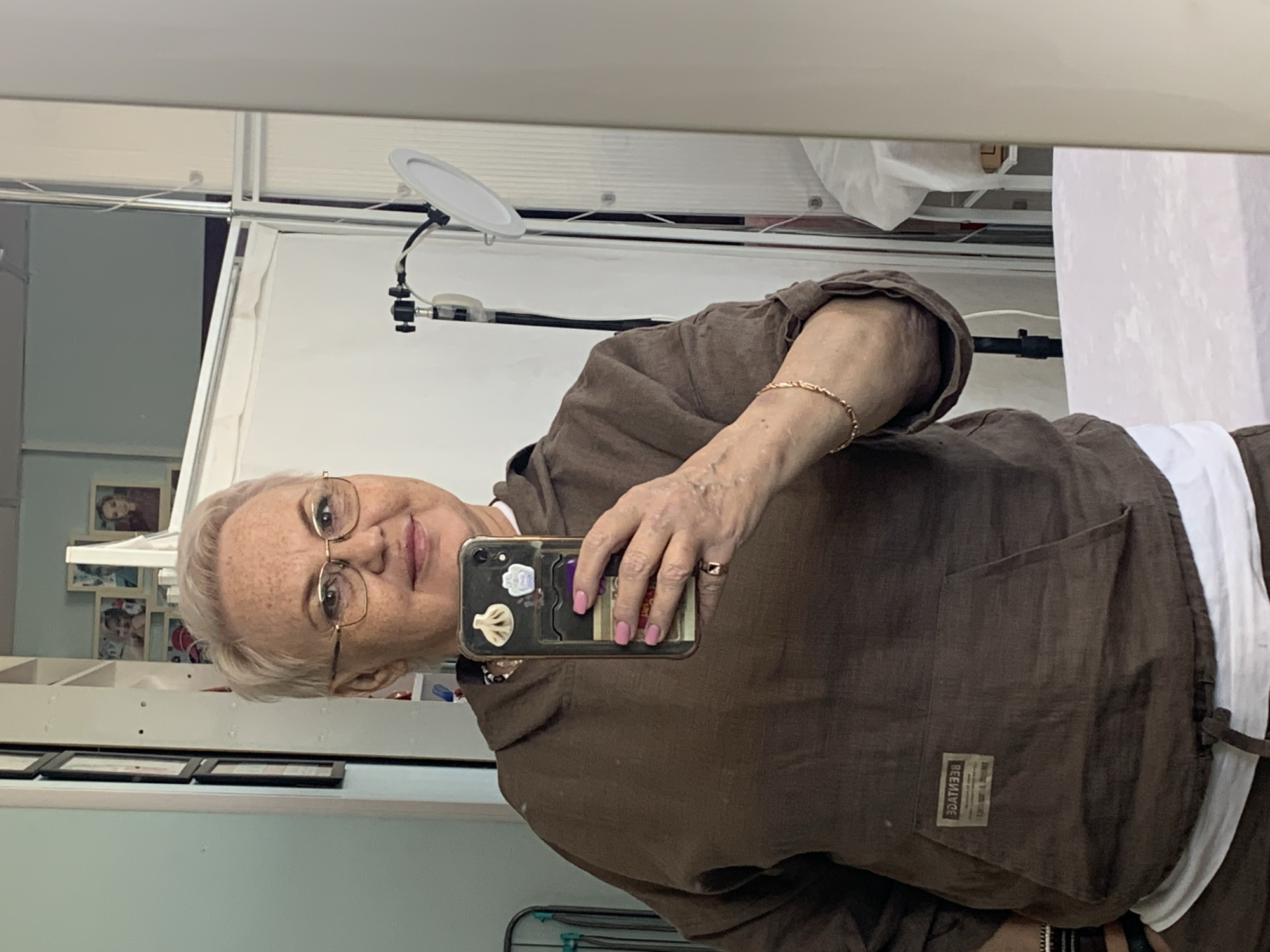 A mirror selfie depicts an older woman with short, gray hair and glasses, wearing a brown hoodie and a white shirt underneath. She is holding a smartphone in her right hand, with pink painted fingernails visible. The background includes a room with light blue walls, white shelves displaying framed photos and other items, and a white backdrop setup with a ring light. The label on the hoodie reads "BEENIVYGE." This image could have been taken in Chaikovsky, Russia, but additional contextual information would be required to confirm its location.