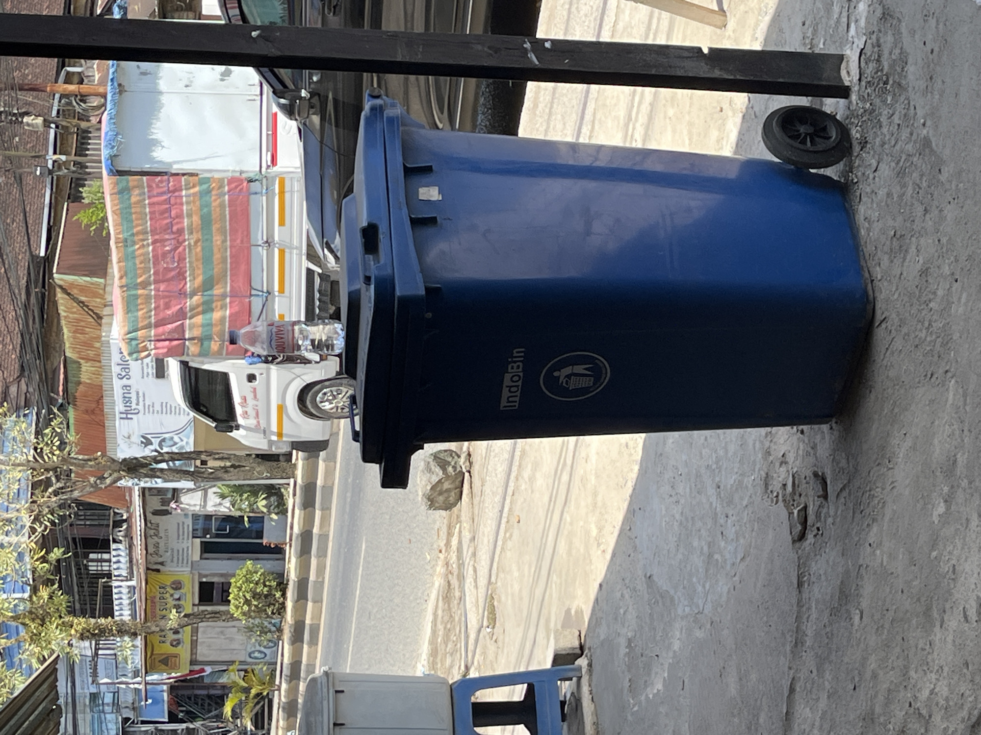 The image depicts a street scene in Sejahtera, Indonesia. In the foreground, a large blue trash bin with wheels stands on a concrete sidewalk. A bottle of Aquavita water is placed on top of the bin. Behind the bin, a black car is parked. Further back, a white truck with a colorful tarp covering its cargo bed is parked on the street. Buildings line the street, some with signs indicating businesses. Trees and foliage are visible, suggesting an urban environment. The overall scene appears to be a typical street view in an Indonesian city.