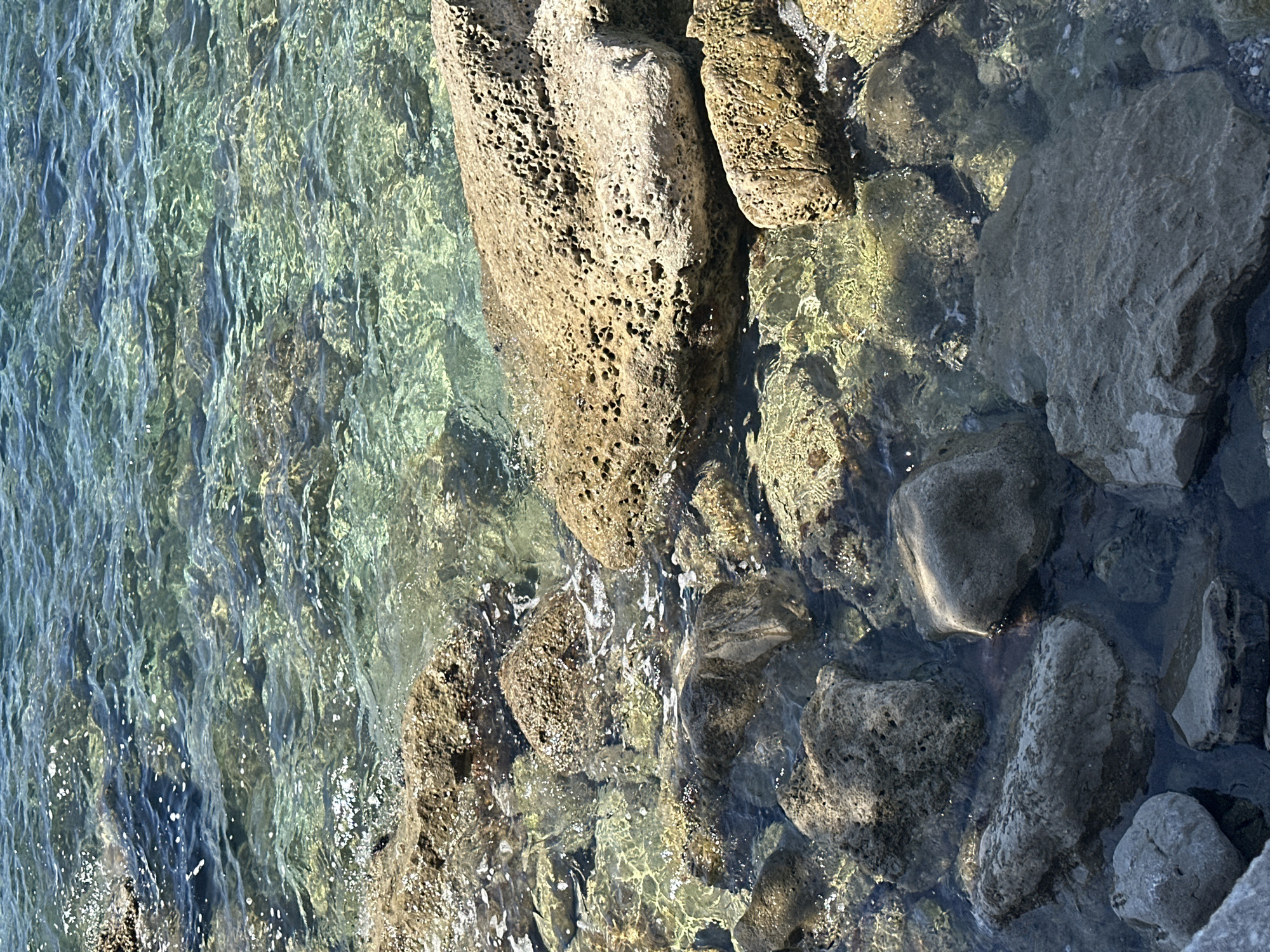 The image shows a shoreline in Budva, Montenegro. Clear, shallow water reveals rocks and stones of varying sizes submerged just below the surface. The rocks are shades of brown and gray and are wet from the sea. The water's surface is rippled, creating patterns of light and shadow. No people or animals are visible.