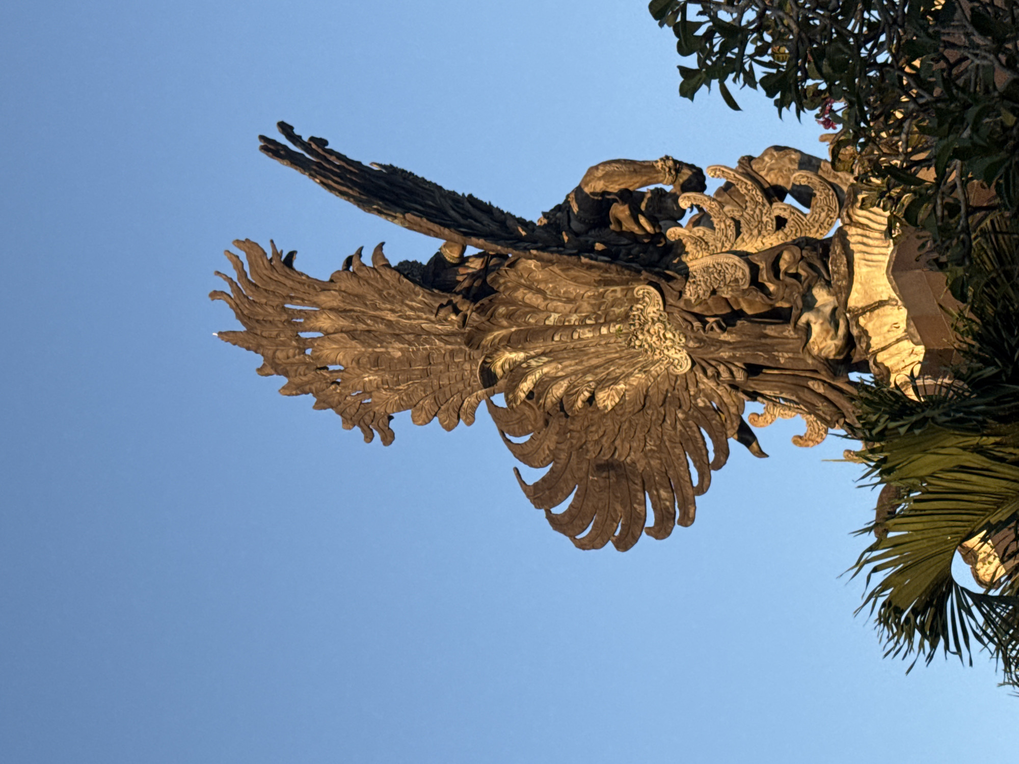 The photograph depicts a low-angle view of a Garuda sculpture against a clear blue sky. The Garuda, a mythical bird-like creature, is rendered in intricate detail with its wings prominently displayed. The sculpture appears to be made of stone or a similar durable material. To the right of the Garuda, foliage from nearby trees is visible, providing a contrast to the smooth, uniform color of the sky. The scene is likely located in Ungasan, Indonesia, given the context provided. The photograph captures the artistic and cultural elements present in the area.