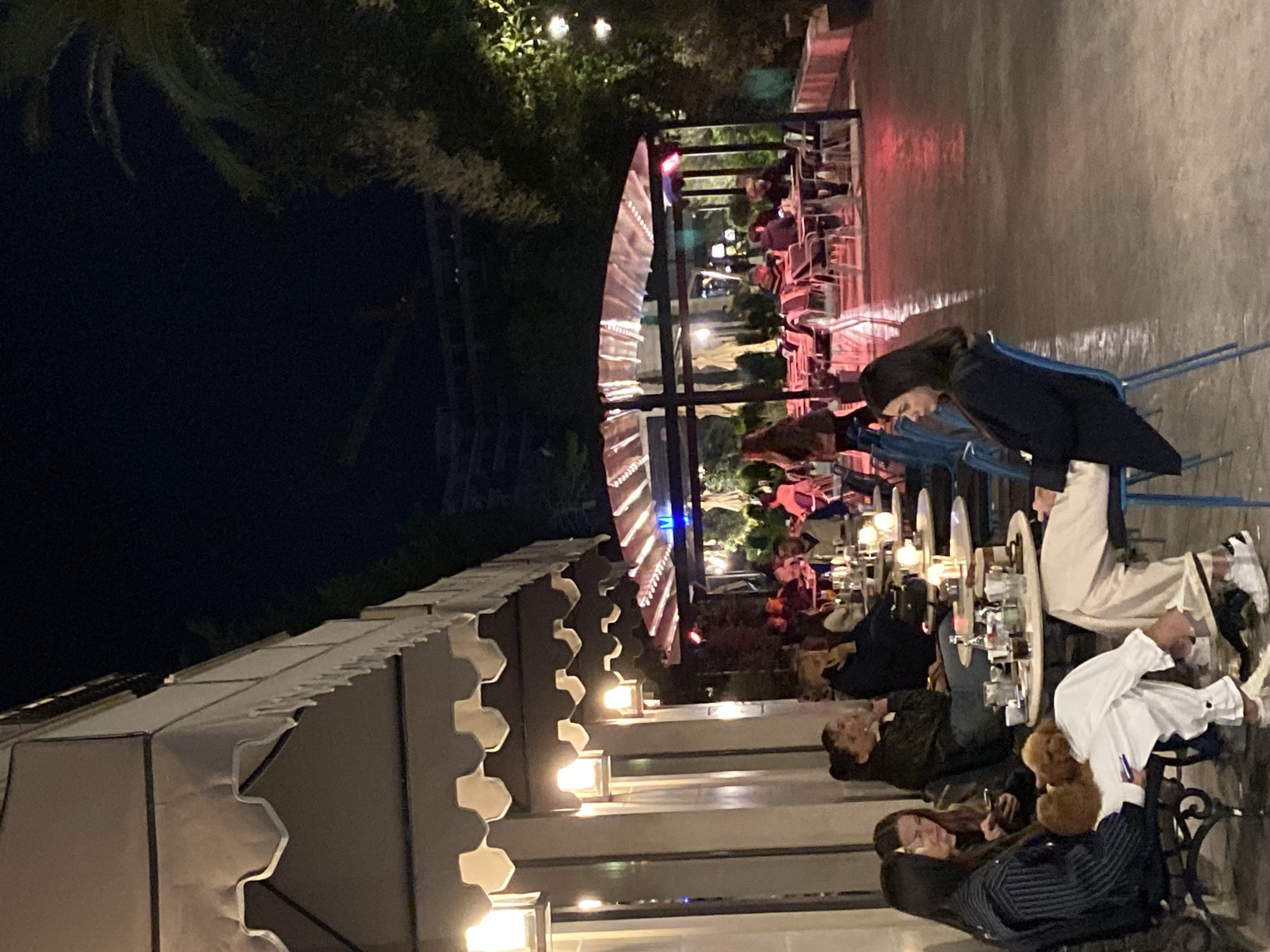 The image depicts an outdoor seating area, possibly a restaurant or cafe, at night. Several people are seated at tables, with drinks and other items visible on the tabletops. The area is illuminated by overhead lights and some lighting fixtures along the building's awning. The background includes greenery and possibly other buildings. Based on the location in Budva, Montenegro, it can be deduced that the seating area is in the open space that is part of the restaurant.