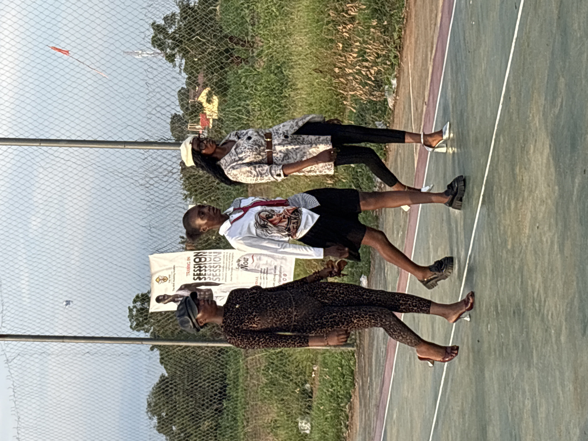 Three individuals are walking on a basketball court in Lafia, Nigeria. The person on the left is wearing a leopard-print catsuit and a black cap, and is walking towards the left. The person in the middle is wearing a white shirt with a graphic print and a black skirt, and is walking towards the left. The person on the right is wearing a patterned jacket, black leggings, and a white hat, and is walking towards the left. The court is green with white lines. In the background, there is a chain-link fence, trees, and a banner that reads "TRAINING IN SESSION". The sky is overcast.
