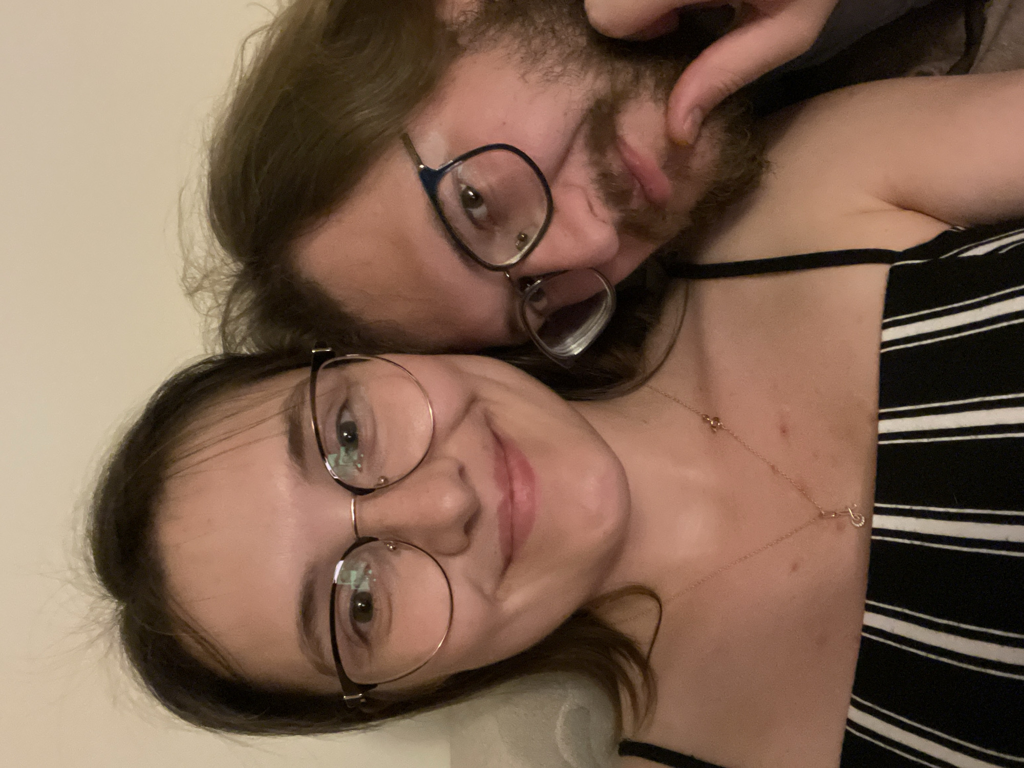 Two individuals are posing closely for a selfie. One person, positioned in the foreground, appears to be a young woman with shoulder-length brown hair, fair skin, and wearing glasses with thin, round frames. She is smiling at the camera and wearing a black and white striped top with thin straps. A delicate gold necklace is visible on her chest.
The second person, likely a male, is behind her. He has long, brown hair, a beard, and also wears glasses. His face is partially obscured. He has fair skin.
The background is plain and appears to be an interior wall, possibly in a residential setting. There is no indication of the location being in Budva, Montenegro.
