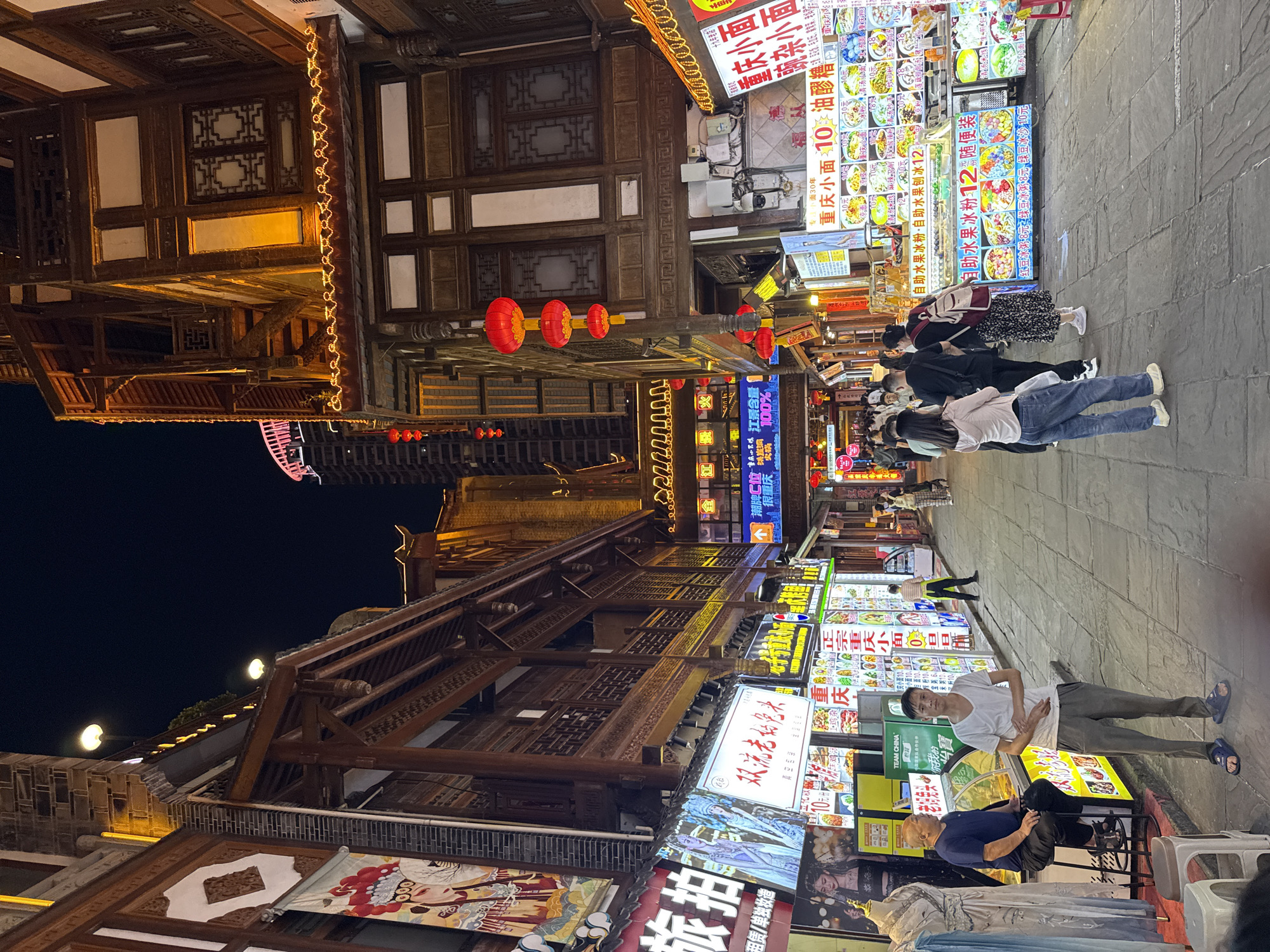 This image shows a bustling street in Yuzhong District, China, at night. The street is lined with traditional Chinese architecture, featuring wooden buildings and red lanterns. Many food stalls are visible, their bright, colorful signs illuminated. People are walking along the street, some stopping to look at the food stalls. The OCR text on the signage shows that various local dishes are being sold. A few individuals are seen standing or sitting near the stalls, possibly customers. The overall scene is lively, suggesting a popular night market or tourist area.