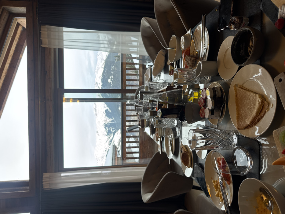 An indoor dining scene within a chalet-style building in Courchevel, France, is captured, presenting a long, dark-surfaced dining table set for breakfast or brunch. The table shows clear signs of recent use, with multiple plates, glasses, and bowls containing food remnants and fresh items. No individuals are present in the image.
The setting is characterized by a large expanse of floor-to-ceiling windows that provide a panoramic view of snow-covered mountains, coniferous trees, and distant chalet-style buildings under a bright, overcast sky. Directly outside the window, a wooden balcony railing is visible. The interior features exposed wooden beams, dark primary curtains pulled to the sides, and sheer white secondary curtains. Several light-toned, upholstered dining chairs are positioned around the table.
On the table, various items include white ceramic plates, some containing remnants of scrambled eggs and bread, while others hold folded crepes. A small platter displays sliced kiwi and raspberries. Numerous small jars of preserves and spreads, including a dark chocolate-like spread in a bowl, are distributed. Clear crystal glasses, several glass bottles of water, a dark bottle of what appears to be oil or vinegar, and a white ceramic creamer jug are present. Cutlery is arranged on and beside the plates. A dark-colored smartphone rests on the table surface.