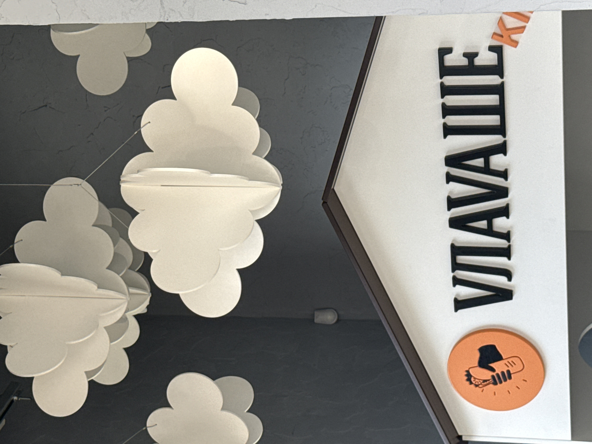 The image shows the exterior of a food establishment, likely a cafe or fast food restaurant, with the name "ВЛАВАШЕ" (VAVASHE) in large black Cyrillic letters on a white facade. To the left of the name is an orange circular logo depicting a hand holding a kebab or shawarma with radiating lines, symbolizing a hot or fresh product. A partial orange letter "К" is visible to the right of the main text. The establishment appears to be a single-story building with a dark brown roofline. Above the facade, several white paper or cardboard cloud-shaped decorations are suspended from the ceiling, creating a whimsical atmosphere. The background wall is dark gray with a textured finish. The image is taken from a slightly low angle, looking up towards the facade and the decorations. The specific location in Lyubertsy, Russia, cannot be determined from the visual information alone.