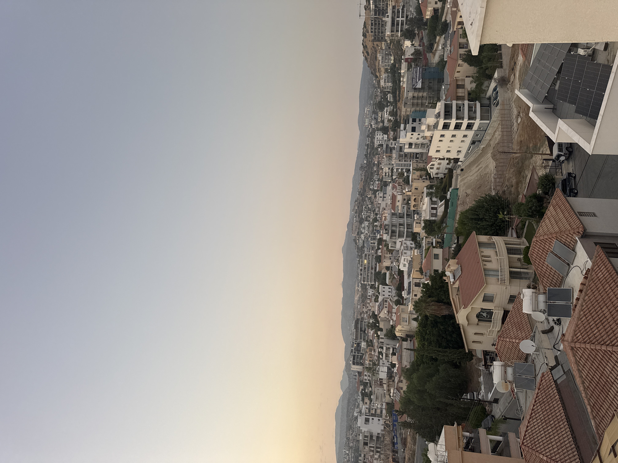 The image captures a wide-angle, elevated view of a residential area in Agios Athanasios Municipality, Cyprus, during what appears to be late afternoon or early evening, given the soft, warm light and the gradient of the sky from pale yellow near the horizon to a light blue overhead.
The foreground is dominated by the tiled roofs of several houses, many of which are equipped with solar panels and water heaters, typical of Mediterranean architecture. Some roofs feature satellite dishes. One prominent house in the mid-foreground has a terracotta-tiled roof, a light-colored facade, and a balcony with decorative railings. Adjacent to it, another house shows a similar roof style.
The middle ground reveals a densely packed urban landscape with a mix of residential buildings and multi-story apartment complexes. Buildings are predominantly white or light-colored, with flat or tiled roofs. Some structures are under construction, indicated by scaffolding and exposed frameworks. There are also areas of exposed earth and dirt roads, suggesting ongoing development. Trees, mostly evergreen, are interspersed throughout the area, providing patches of green against the urban sprawl.
In the background, a range of undulating hills forms the horizon, their slopes dotted with vegetation and some scattered buildings. The overall impression is a thriving, developing suburban or semi-urban environment, likely in a hilly region of Cyprus. No people are clearly visible in the frame, but several vehicles, including cars and what appears to be a construction vehicle, are parked on streets or in driveways. The scene conveys a sense of everyday life in a sun-drenched locale.