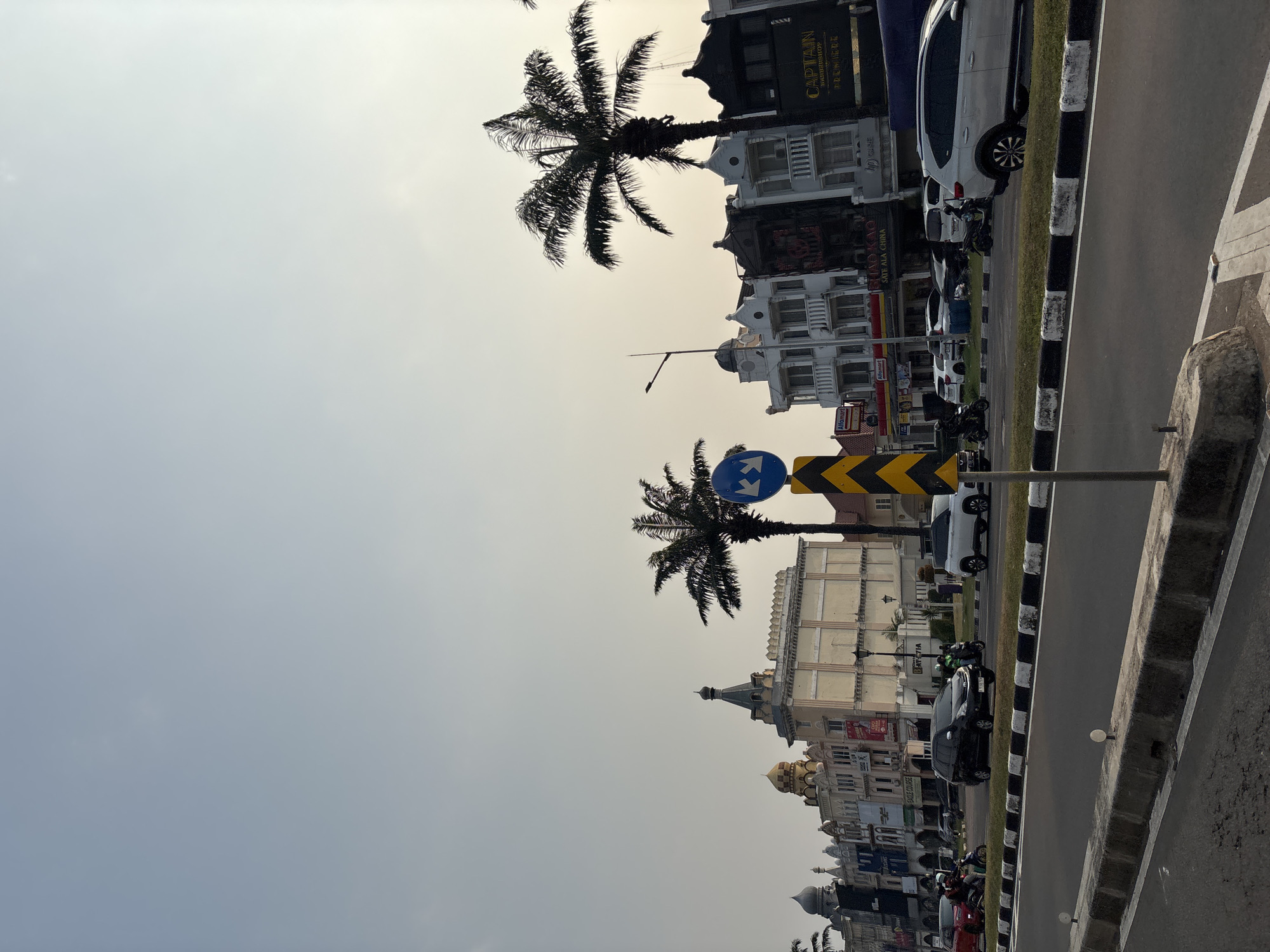 The image captures a street scene in Pakulonan Barat, Indonesia, with a row of multi-story buildings lining the right side of a road. The buildings exhibit diverse architectural styles, including ornate facades with balconies and what appear to be decorative domes and spires. Palm trees are interspersed among the buildings. Several vehicles, including cars and motorcycles, are present on the road, with some in motion. A traffic sign with blue and white symbols is visible on a pole. The sky is overcast, suggesting it could be morning or late afternoon. The foreground shows a paved road with a concrete barrier and grass median. There are no clearly identifiable people in focus, though movement suggests activity. The overall impression is of a developed urban commercial area.