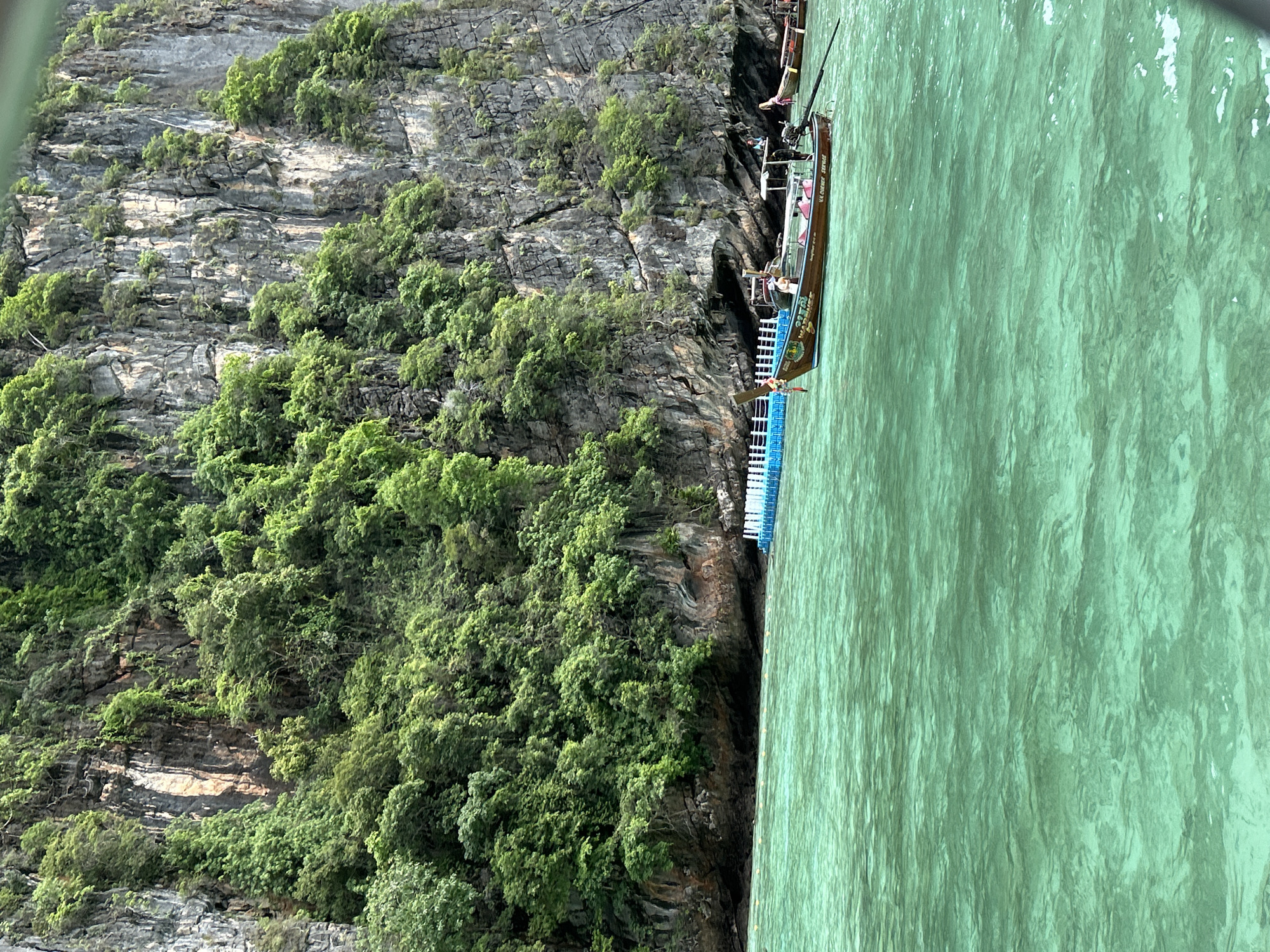 In Ao Nang, Thailand, a longtail boat is anchored in the emerald waters of the Andaman Sea, near a towering limestone cliff face densely covered in green foliage. The boat, with its distinctive wooden hull and protruding propeller shaft, has two people visible on board. In the distance, a floating platform with a white railing is partially submerged, and another longtail boat is visible to the right. The water exhibits ripples and variations in color, suggesting shallow depths in some areas. The overall scene captures a serene tropical seascape.