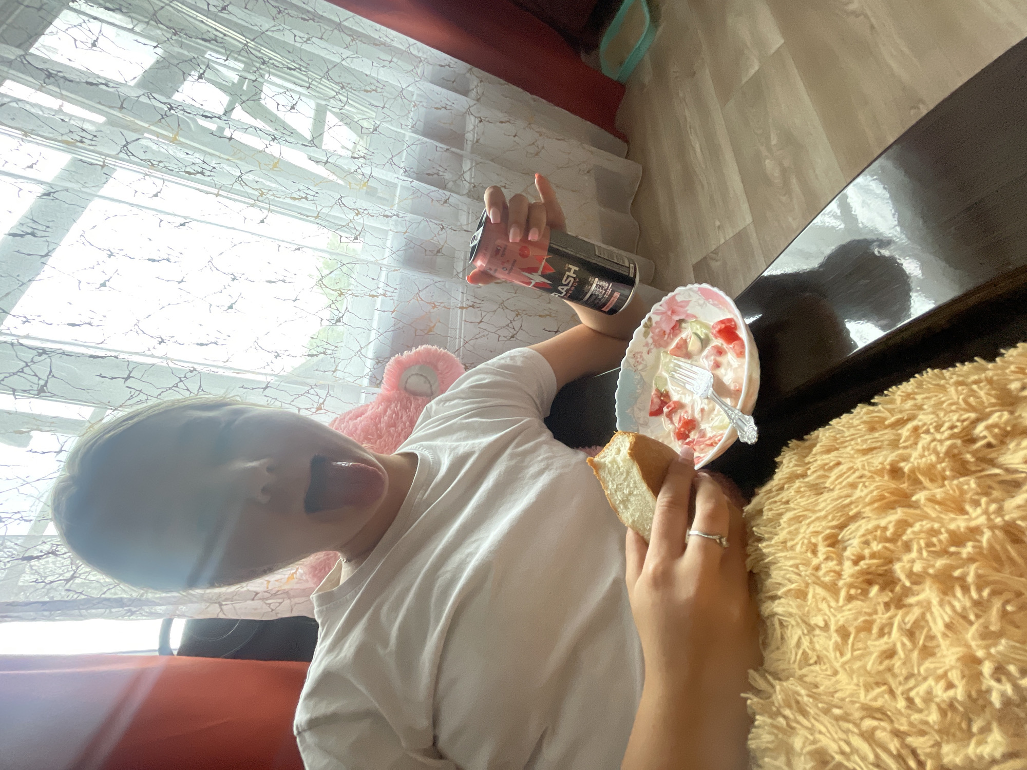 In a residential setting in Медведево, Russia, a young person is seated indoors. They are wearing a white t-shirt and have their tongue sticking out, with their eyes closed. In their right hand, they are holding a can of "FLASH ENERGY" drink. Their left hand is holding a piece of bread, and they are positioned near a plate containing what appears to be the remnants of a meal, possibly a salad with tomatoes and other ingredients. The background shows sunlight streaming through a window with sheer curtains. A fuzzy, light-colored blanket is visible in the foreground. The overall scene depicts a casual moment of eating and drinking.