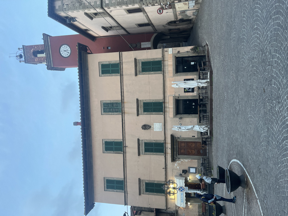 The image displays a central piazza in Castel del Piano, Italy, under an overcast, light blue-gray sky. The foreground is paved with dark gray cobblestones arranged in a fan pattern, featuring white circular markings and four dark, large planters, some containing foliage. Two individuals are present on the lower left: an adult in a blue jacket facing right, and a child in light clothing holding a light blue object, facing the adult near the planters.
The dominant structure is a three-story, light peach-colored building with a dark roofline. It features multiple rectangular windows with dark green wooden shutters, stone sills, and surrounds. A decorative stone crest and a rectangular plaque are affixed to the second story. The ground floor houses a "RISTORANTE" on the left, identified by a white awning and an ornate lamppost. A large, dark wooden double door is centrally located, flanked by two dark-fronted commercial establishments to its right. Numerous dark tables and chairs are arranged for outdoor dining, accompanied by two large, white, folded patio umbrellas.
To the right and slightly behind this building, a tall, square, red/terracotta clock tower rises. Its circular clock face, marked with Roman numerals, indicates a time of approximately 9:55. The tower culminates in an open belfry structure housing a bell and a small cross. An arched stone passage extends from the right side of the main building, leading to a narrow, uphill cobblestone street (Via Borgo) that ascends past another multi-story building constructed of lighter stone masonry, featuring a "No Entry" sign on its wall.