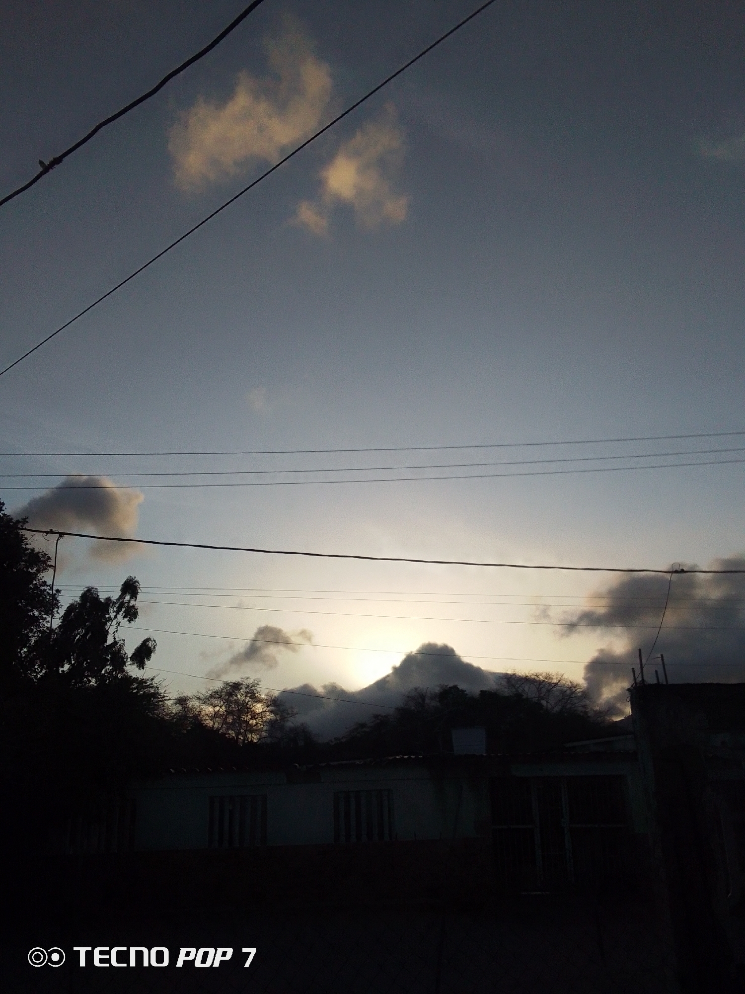 The image captures a view in Altagracia, Venezuela, at dusk or dawn. The sky is predominantly a muted blue-gray, with a few scattered clouds tinted with hints of yellow from the rising or setting sun. Several power lines cut across the frame. Below the horizon, a silhouetted mountain range is visible, obscured by trees and a building. The building appears to be a single-story structure with windows. In the foreground, the scene is dark. The camera used is identified by the watermark "Tecno Pop 7."