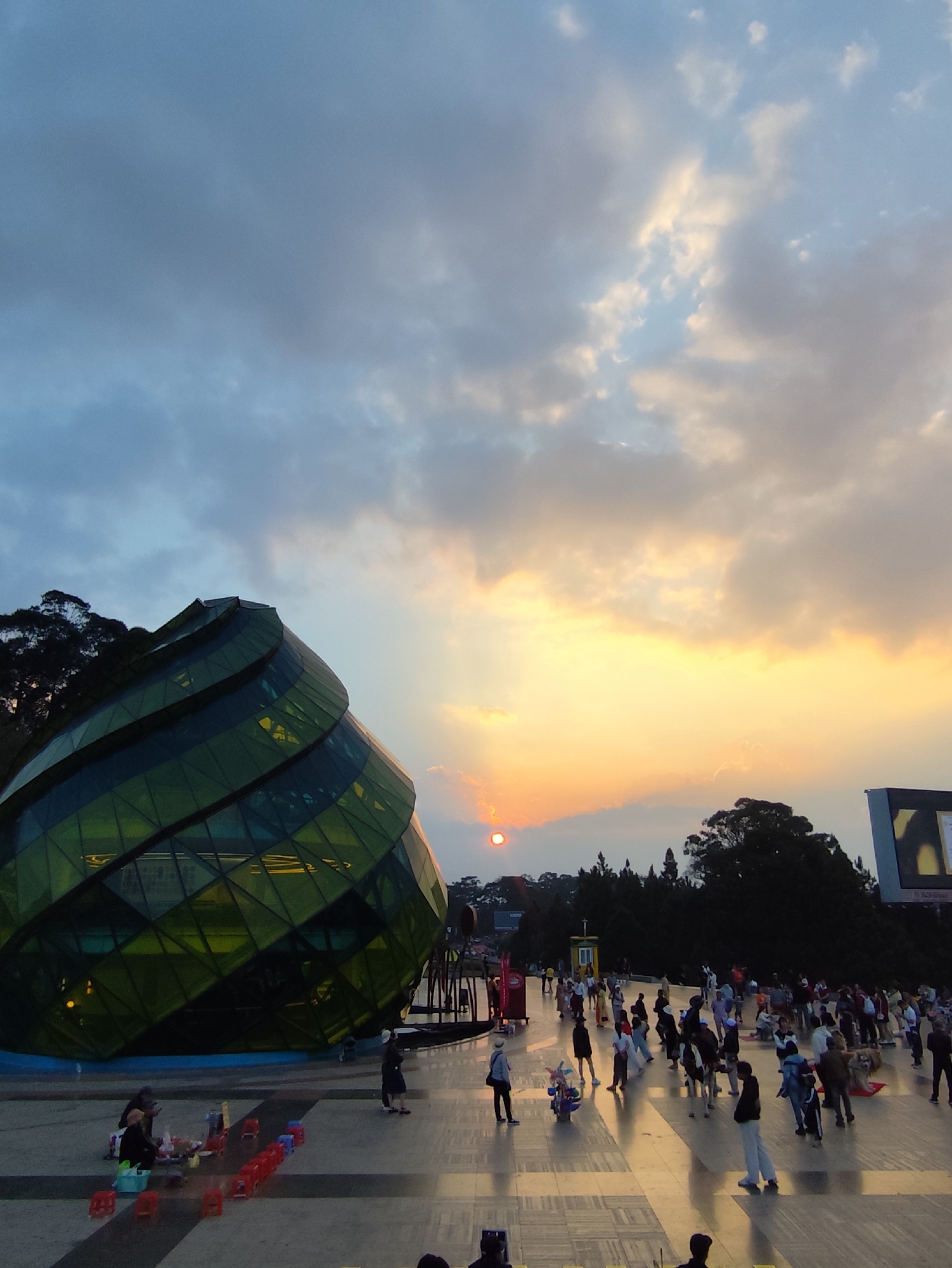 The photo shows a bustling public square in Da Lat, Vietnam, at sunset. The prominent structure is the glass-covered, cone-shaped building of Square Lam Vien, a recognizable landmark in the city. Numerous people are milling about the square, some appearing to be tourists. Vendors are selling goods, with a small setup of plastic stools and items displayed on the ground visible on the left. The sky is filled with clouds tinged with orange and yellow hues from the setting sun. Other buildings and trees are visible in the background.