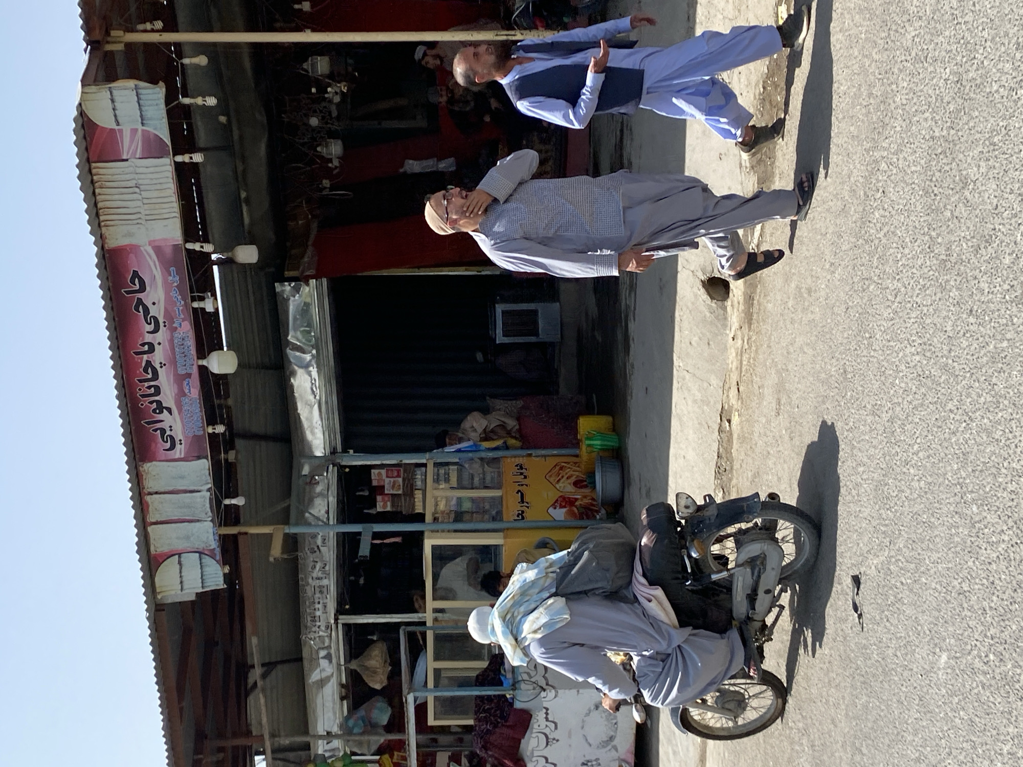 In the city of Kandahar, Afghanistan, a man on a motorcycle rides away from the camera, wearing a light blue and white checkered cloth draped over his shoulders and a grey garment. He carries a dark grey bag on the back of his motorcycle. Two men walk on the right side of the road, towards the motorcycle. The man closer to the camera wears a light blue vest over a light blue tunic and pants, with dark shoes. The man further away wears a grey patterned tunic and grey pants, with dark sandals. They are walking on a paved road in front of a market stall. The market stall has a corrugated metal roof and displays various goods. The background shows a clear blue sky and a simple building structure.