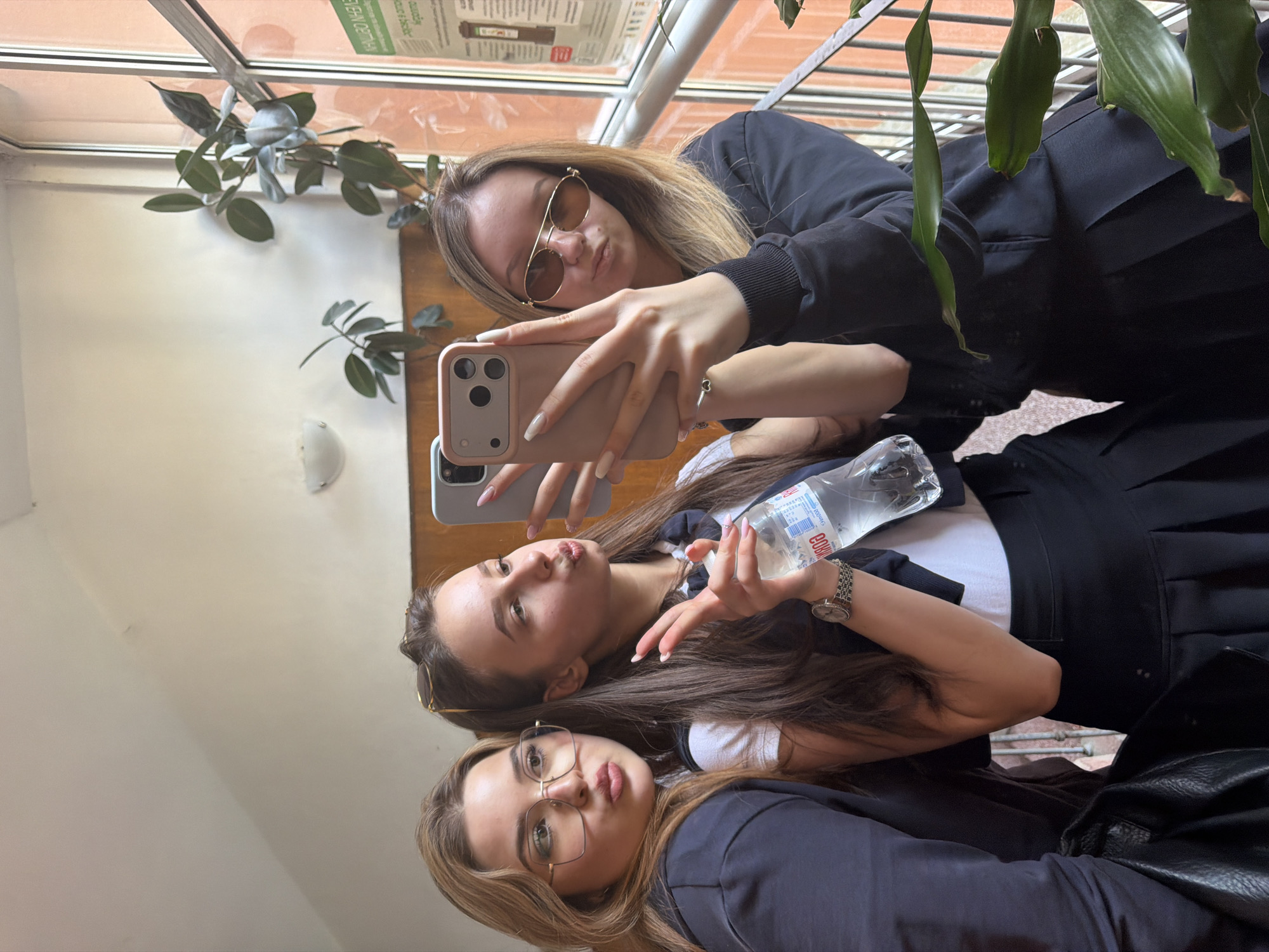 Three young women are taking a selfie indoors. The woman in the top of the frame is holding a pink smartphone. All three are pouting their lips. They are wearing glasses. One of them is holding a bottle of water. There are plants in the background, and a window. The location is undetermined but potentially in Blagoevgrad, Bulgaria.