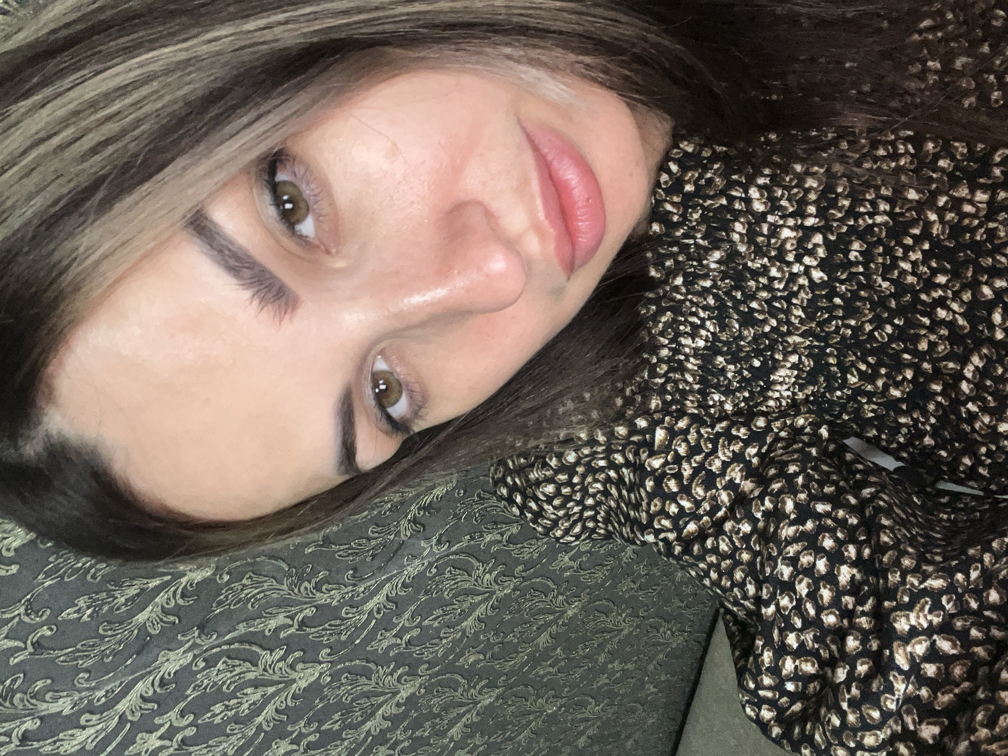 A close-up, eye-level, indoor selfie captures a woman with long, brown, highlighted hair and brown eyes. She is looking towards the camera, her head tilted slightly to the right. Her makeup is subtle, with defined eyebrows and a natural lip color. She is wearing a dark, patterned top with a smock-like texture. The background features a textured wallpaper with a repeating floral or leaf motif in shades of dark gray and gold. The lighting appears to be soft, highlighting her facial features and the texture of her clothing and the background. The overall setting suggests a casual, indoor environment.