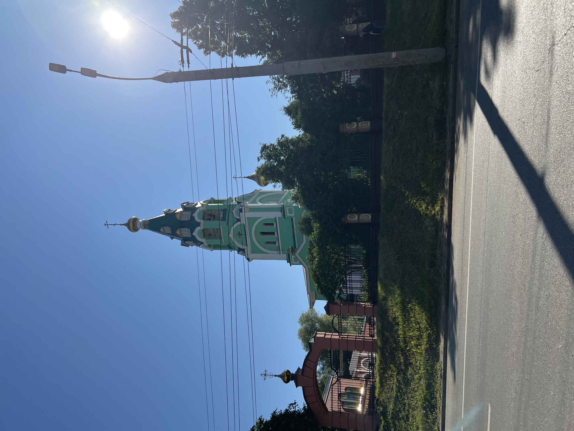 The image captures the Holy Trinity Cathedral in Izhevsk, Russia, under a clear blue sky. The cathedral's green and white architecture, including its tall bell tower topped with a golden cross, is prominent. A red arched gateway with a smaller golden cross stands to the left, leading into the church grounds, which are enclosed by a dark fence and lush greenery. A lamppost with two lights is visible on the right, casting a long shadow across the asphalt road in the foreground. The sun is shining brightly, creating lens flare in the upper right corner and illuminating the scene. A person in dark clothing is partially visible walking near the fence on the right side of the image. The overall scene depicts a peaceful day in a city.
