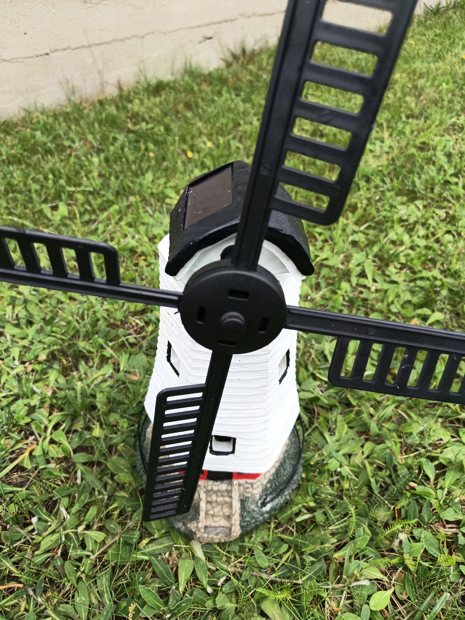 This is a close-up, eye-level, outdoor, full shot of a decorative windmill ornament. The windmill is white with black blades and a black roof. It has a solar panel on top. The base of the windmill is grey and green, with a small stone staircase leading up to it. The windmill is positioned on a patch of green grass with some small yellow flowers. In the background, a light-colored concrete wall is visible. The scene is likely located in the city of Лосева, Belarus, given the provided information. There are no people or animals present in the image.