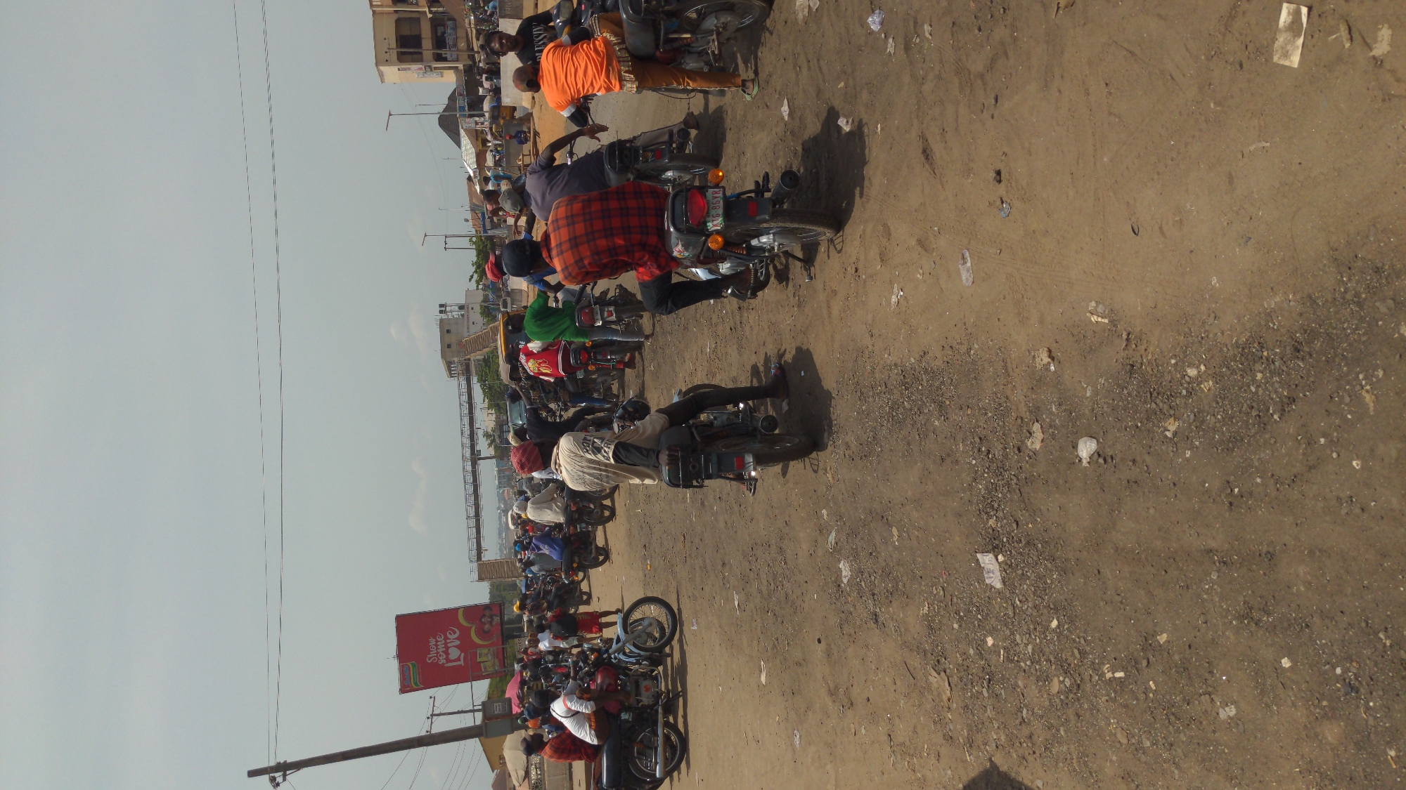 This image captures a busy, unpaved street scene, likely in an urban or semi-urban area, under bright daylight conditions with a clear sky. The time of day appears to be mid-morning or afternoon, judging by the strong shadows cast by people and vehicles.
The primary subjects are numerous people on motorcycles, dominating the foreground and midground. Most individuals are seen from behind, riding or waiting on their bikes. They are dressed in casual attire, with some wearing hats or caps. Many of the motorcycles appear to be "okadas" (motorcycle taxis), a common mode of transport in Nigeria. The ground is a dusty, reddish-brown dirt road, showing tire tracks and mixed with gravel and litter, including plastic bottles and pieces of paper.
Along the left side of the road, in the background, are several multi-story buildings of varying construction styles. Overhead power lines are visible against the pale sky, stretched between poles. A large, red billboard is positioned on the left side of the road in the midground. The most prominent text on the billboard reads "SHOW SOME LOVE," accompanied by other smaller, less legible text and graphic elements. One visible motorcycle license plate clearly displays "LAG 556 RR." The scene depicts a lively local thoroughfare characterized by significant motorcycle traffic and a dusty environment.