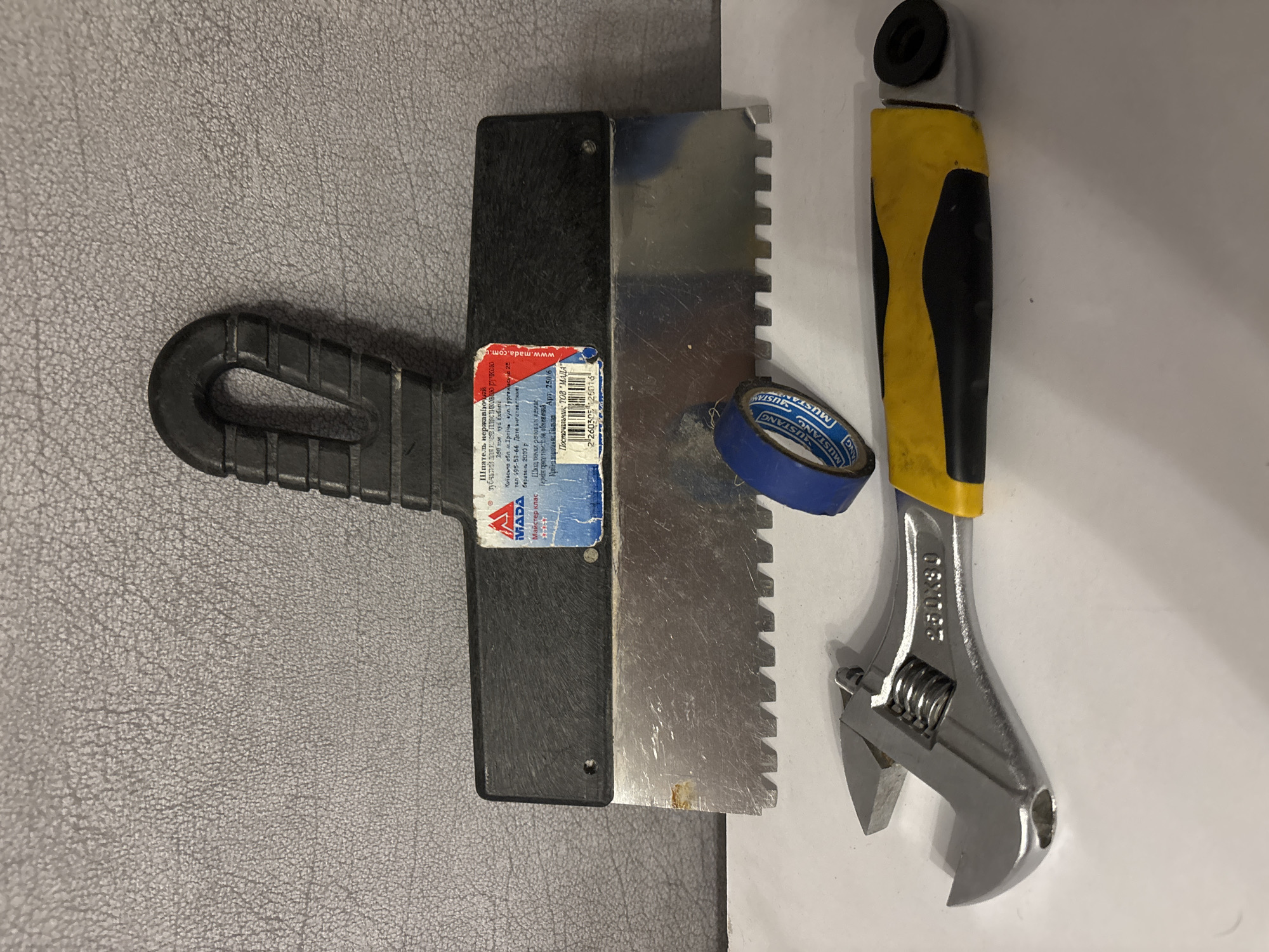 A notched trowel, a roll of blue electrical tape, and an adjustable wrench are arranged in a still life against a textured gray background. The trowel, positioned at the top, has a black plastic handle and a stainless steel blade with serrated edges. A label is affixed to the trowel's handle, with text in Cyrillic script. To the right of the trowel, a partially used roll of blue tape is centered. Below these items, an adjustable wrench with a silver metal head and a black and yellow rubberized handle lies horizontally. The wrench displays the marking "250x30." The background is a flat, gray surface with a subtle, pebbled texture. The lighting is even, casting no significant shadows. The objects are clean and appear to be in good condition. No people are present in the image. The scene is indoors.