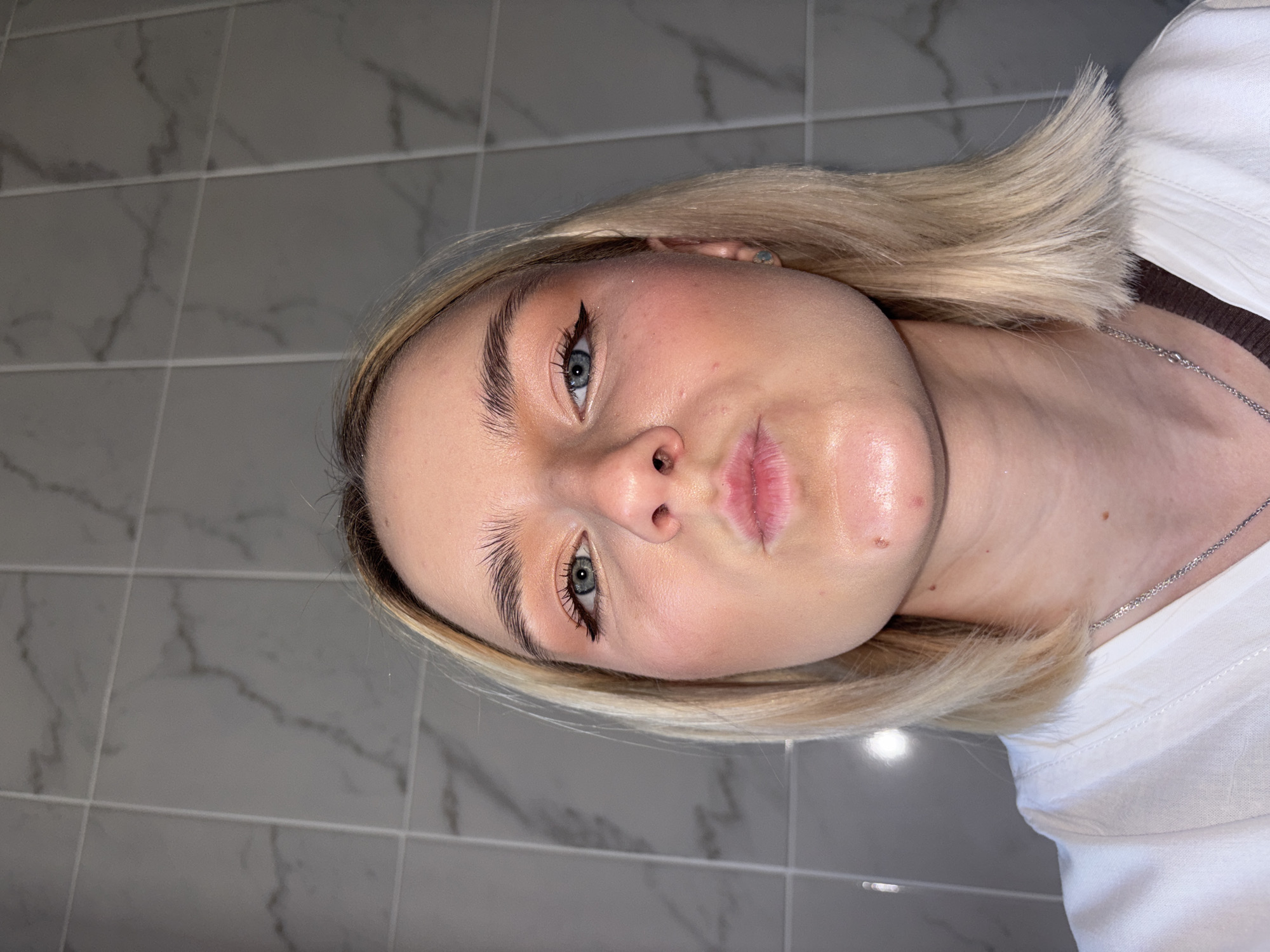 A young woman with fair skin, blonde hair, and blue eyes is the central subject of this close-up portrait. The image is rotated 90 degrees clockwise, showing her head tilted towards the right. She looks directly at the camera with a neutral expression. Her makeup includes prominent winged black eyeliner and neatly styled, dark eyebrows. Her lips are slightly parted and appear glossy, and her skin has a dewy finish, reflecting light particularly on her forehead and around her mouth. Small moles are visible on her neck and near her mouth.
She is wearing a white t-shirt and a thin silver chain necklace. A dark brown garment is visible beneath her white shirt on her left side. Her blonde hair is styled in a short, straight bob or lob cut, framing her face.
The background consists of a tiled wall, characteristic of an indoor setting, possibly a bathroom. The tiles are square or rectangular, featuring a grey and white marble-like pattern with light-colored grout lines. The lighting appears to be artificial, highlighting certain areas of her face and skin. There is no discernible activity taking place other than posing for a photograph. No text is visible in the image. The image itself does not contain visual cues to indicate the location of Saint Petersburg, Russia.