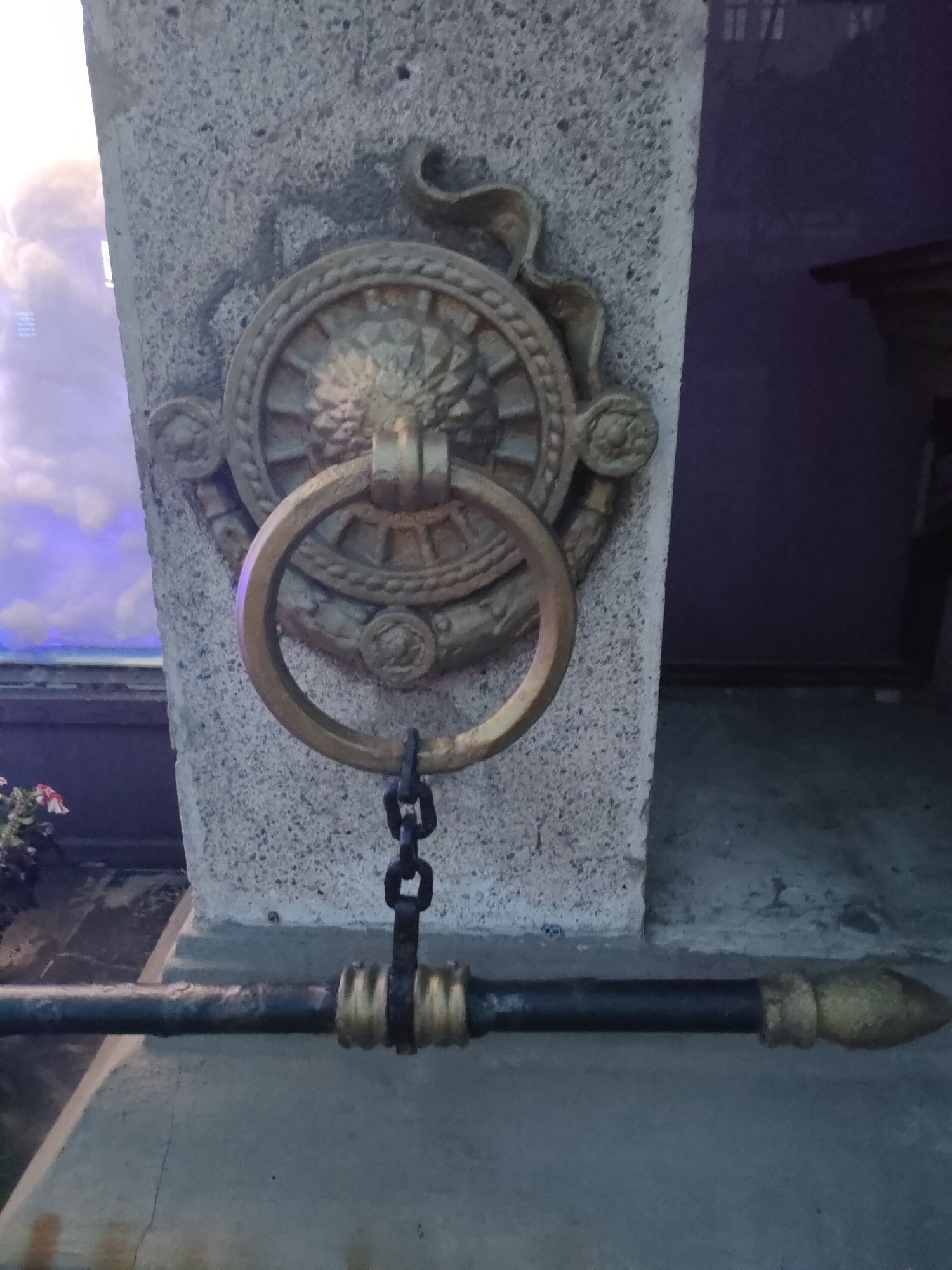 The image shows a close-up of a decorative brass ring and chain attached to a stone pillar. The ring is ornate with a central wheel-like design. Below the ring, a black metal bar with decorative brass accents runs horizontally across the frame. The bar has a pointed finial on the right end. The scene is lit with artificial light, casting shadows. The background is dark and out of focus, with hints of a purple wall and a glass pane reflecting light and sky. There are no people or animals visible. The location is not specified in the image or context.