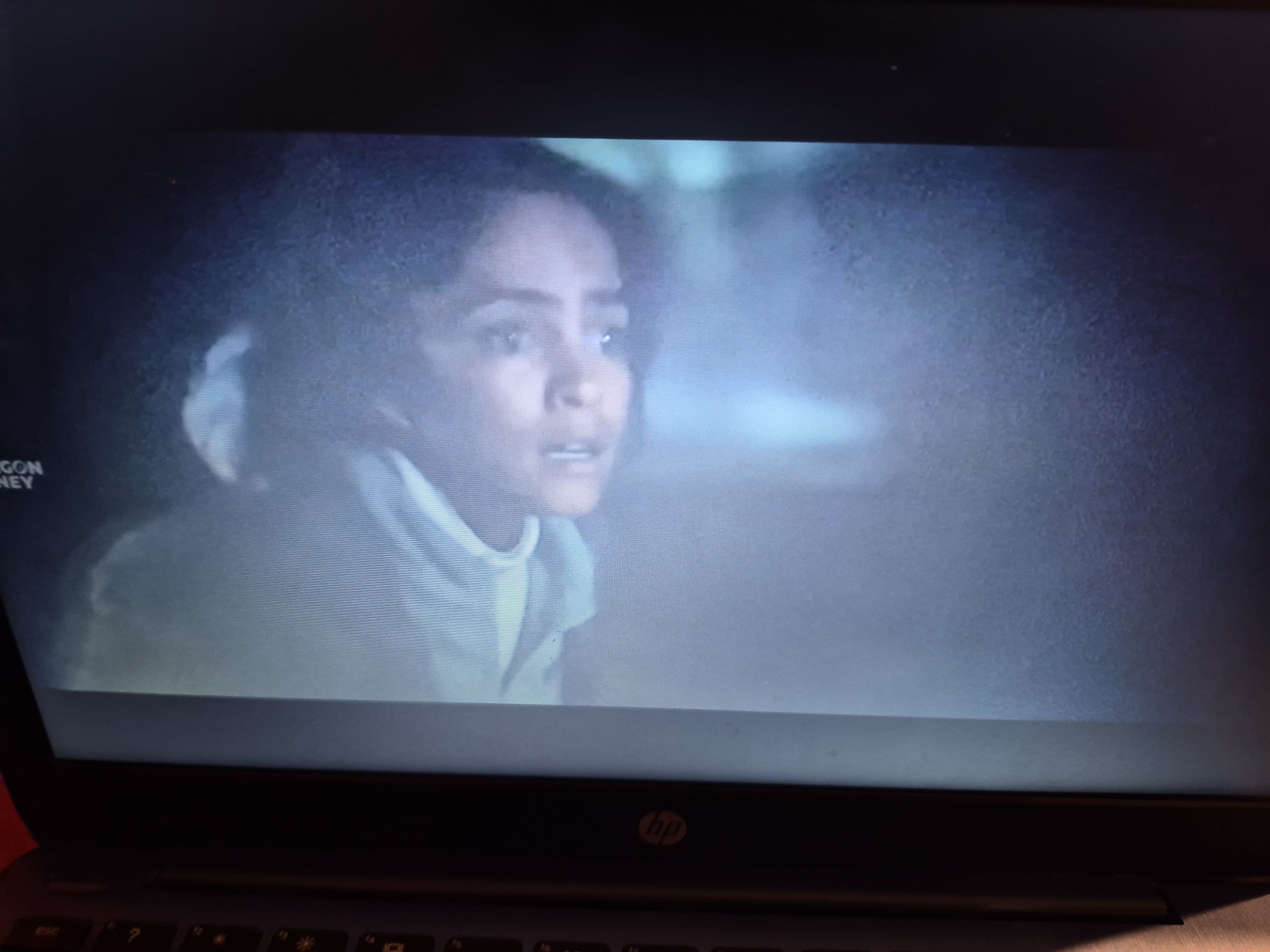 The image shows a close-up, slightly blurry view of a young woman's face on a laptop screen. She has dark, curly hair and appears to be looking to her left with a concerned or surprised expression. She is wearing a light-colored, high-necked garment. The background on the screen is a muted, blue-grey hue, suggesting a dim or atmospheric setting. The bottom of the image shows the keyboard and the HP logo of the laptop. There is text visible in the top left corner of the screen, which reads "GON NEY". The location is not identifiable in this image; it is a generic scene displayed on a laptop.