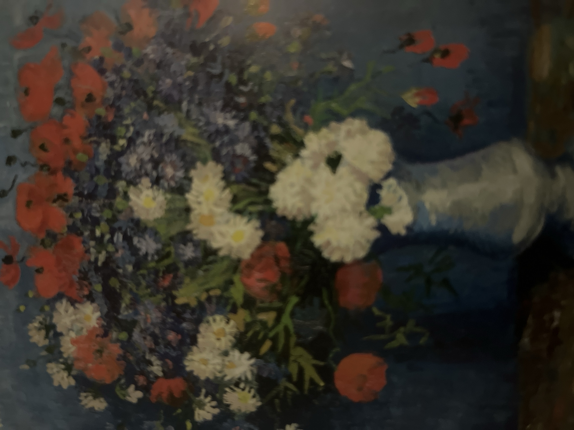The image is a painting of a floral arrangement in a vase against a blue background. The arrangement consists of various flowers, including red poppies, white daisies, and small blue flowers. The vase appears to be light blue or gray and has a rounded shape. The painting is done in an impressionistic style, with visible brushstrokes and a focus on color and light. The scene is set indoors, likely on a table or surface, as suggested by the hint of a darker surface at the bottom of the frame. There are no people visible in the painting, nor any indication of the city of Edem, Russia.