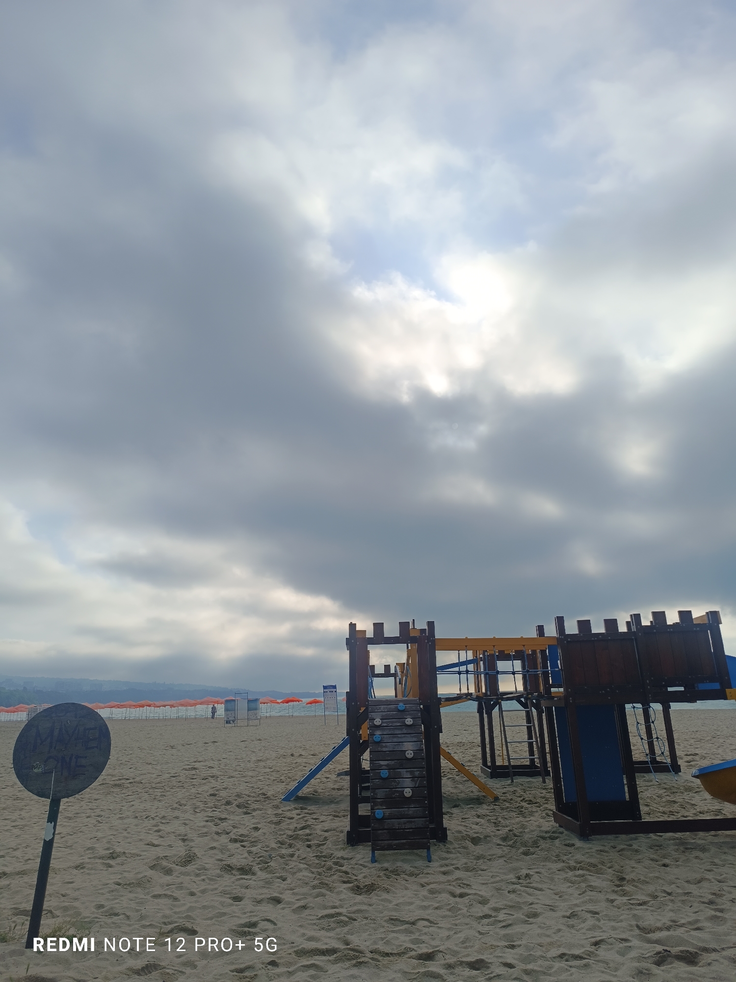 A wide-angle photograph captures a quiet, cloudy day at a sandy beach in Varna, Bulgaria. The foreground features a multi-component wooden playground structure on the right, resembling a small fort or castle, complete with steps, climbing elements, and a blue slide. To the left, a dark, circular sign on a pole displays faded white text that appears to read "MAYBE ONE". The sandy ground is textured with numerous footprints and disturbances.
In the midground, the beach stretches into the distance, lined with rows of closed, orange beach umbrellas. A single, indistinct figure can be seen far away near the water's edge among the umbrellas. Beyond the beach and umbrellas, the calm sea meets a hazy distant coastline, where faint buildings are visible, suggesting a city or town.
The sky is heavily overcast with thick, grey clouds, though patches of brighter sky and obscured sunlight are visible breaking through, particularly in the upper right quadrant. The subdued lighting suggests a cloudy daytime, possibly early morning or late afternoon. The scene conveys a sense of tranquility and anticipation, with the empty playground and closed umbrellas hinting at warmer, busier periods. No specific activity is taking place. A "REDMI NOTE 12 PRO+ 5G" watermark is present in the bottom left corner of the image.