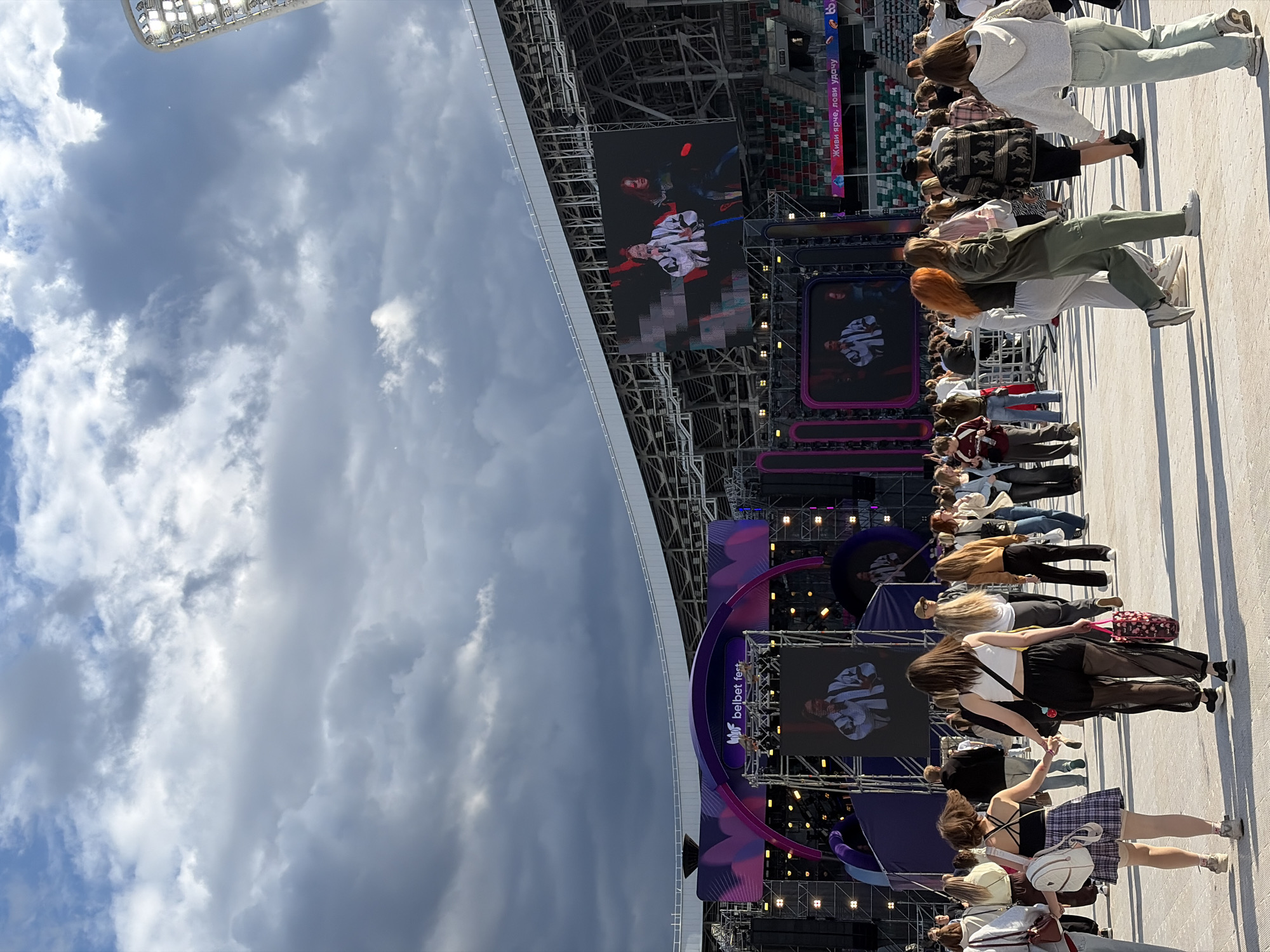 This is a medium-shot, eye-level photograph of a crowd of young people at an outdoor music festival. The festival appears to be held in a stadium, with a large stage set up in the background. The stage has a large video screen displaying performers, as well as elaborate lighting structures. The crowd is composed of many individuals, mostly young adults, dressed in casual, fashionable clothing. Many are walking, some in small groups, while others are holding hands or walking alone. The overall atmosphere suggests a lively and energetic event. The lighting and the presence of a stadium indicate it's likely daytime, with some cloud cover visible in the sky. The logo on the stage reads "belbet fest." The crowd and stage setup are consistent with a music festival taking place in Minsk, Belarus, possibly at the Minsk National Olympic Stadium, which is a common venue for large-scale events.