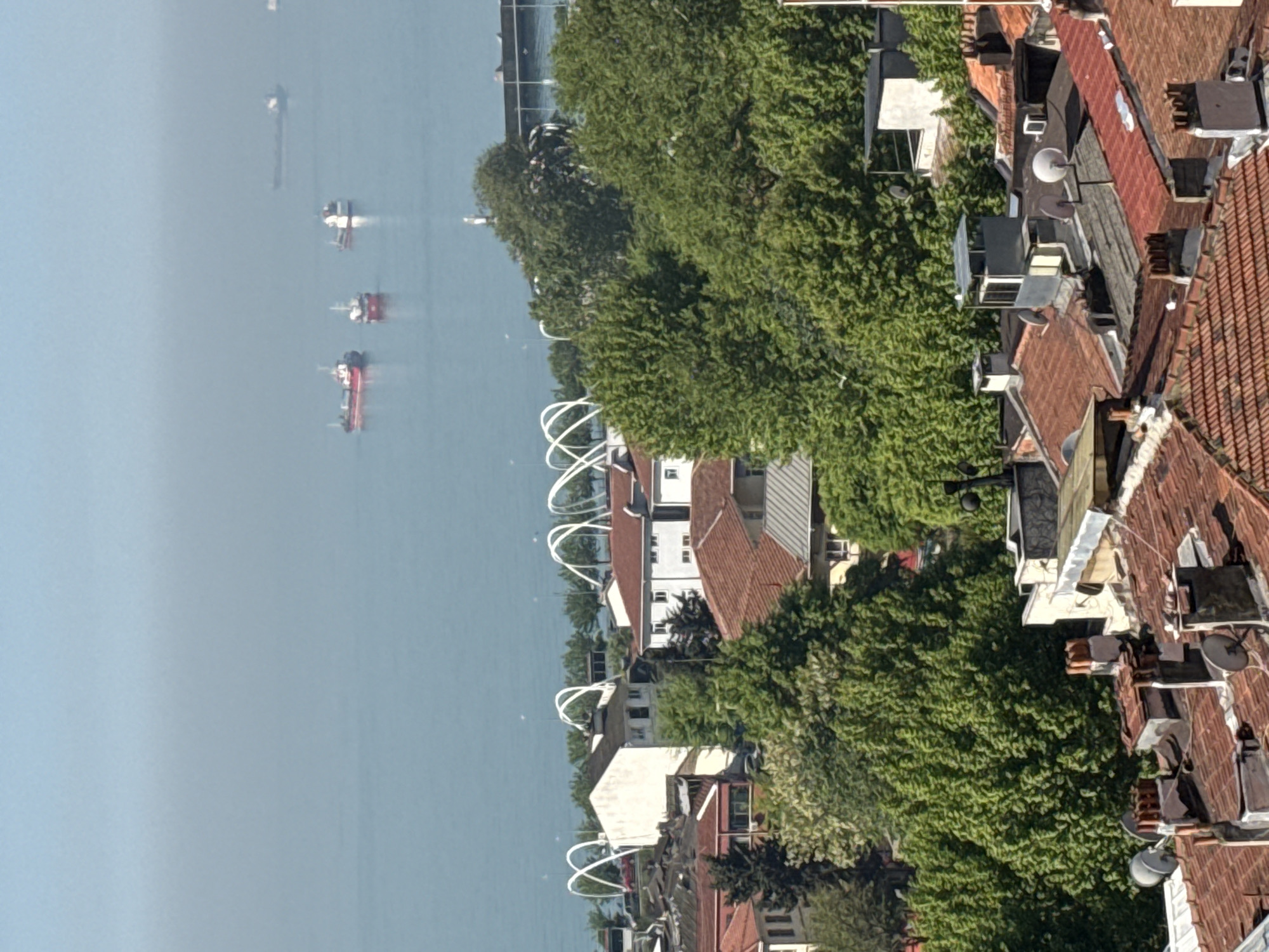 Here is a detailed description of the image:
The image presents an elevated, possibly rooftop view of a coastal urban area. In the foreground and right side, there is a cluster of buildings with red-tiled roofs, various chimneys, satellite dishes, and other rooftop infrastructure. These buildings are interspersed with dense green trees and foliage.
Moving towards the center of the image, distinctive white architectural arches are visible, seemingly part of a waterfront promenade or park. Beyond these arches is a body of water, likely a sea or a bay, which occupies a significant portion of the left side of the frame.
In the water, there are several ships of different sizes. The water appears calm with a slight haze, suggesting a somewhat overcast or misty day.
The location appears to be a city with a coastline. The style of architecture with the red-tiled roofs is characteristic of many European coastal cities. The overall composition suggests a blend of urban density with natural elements like trees and water.