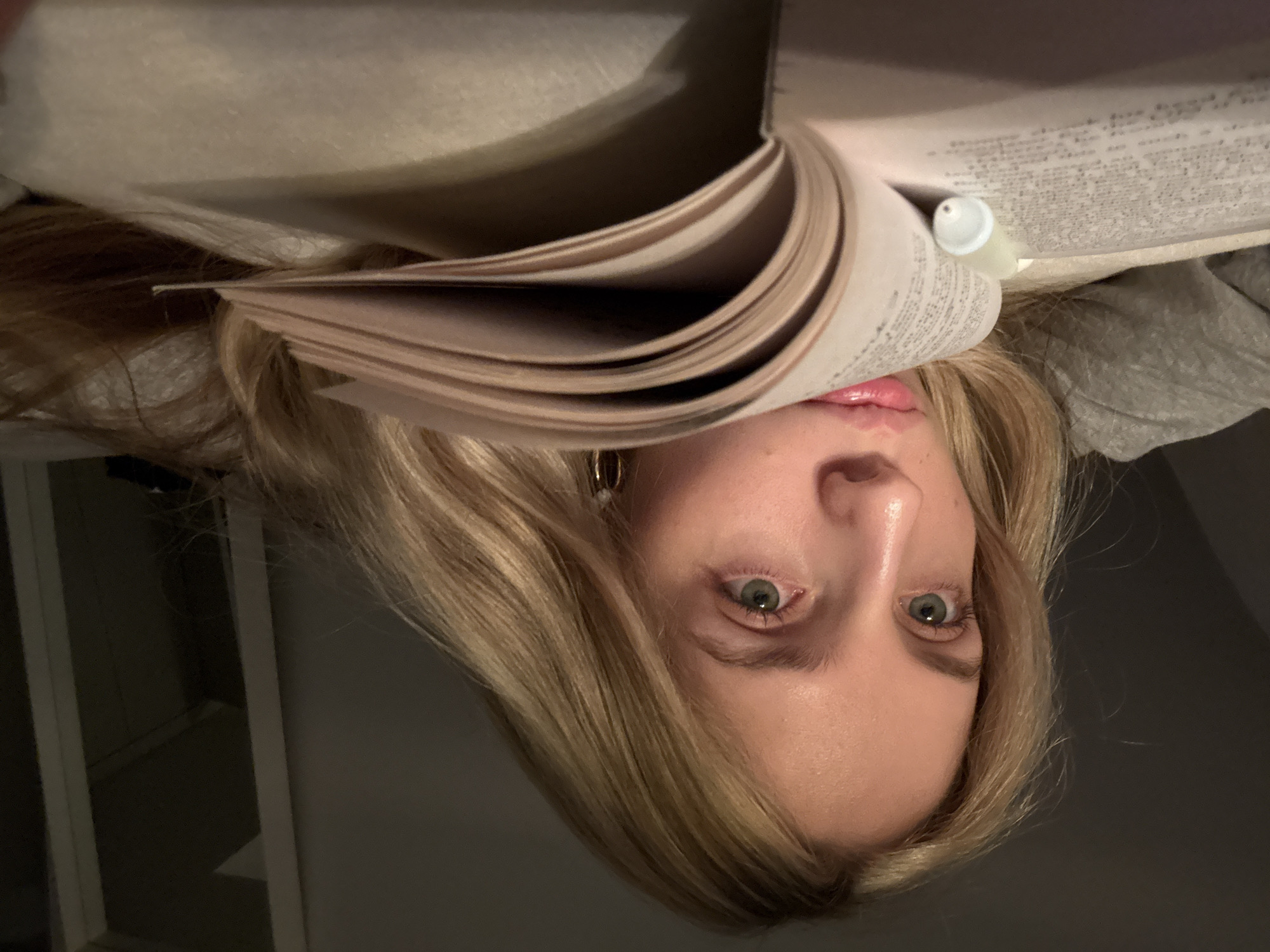 A young, fair-skinned woman with blonde hair is upside down, positioned below an open book. The book is held up at an angle, with some text visible on the pages. She has light green eyes and is wearing a gold hoop earring. The background is blurred, with the corner of a window visible in the lower left and a dark area on the right. A small tube is wedged in the book's spine. The location is not discernible from the image.