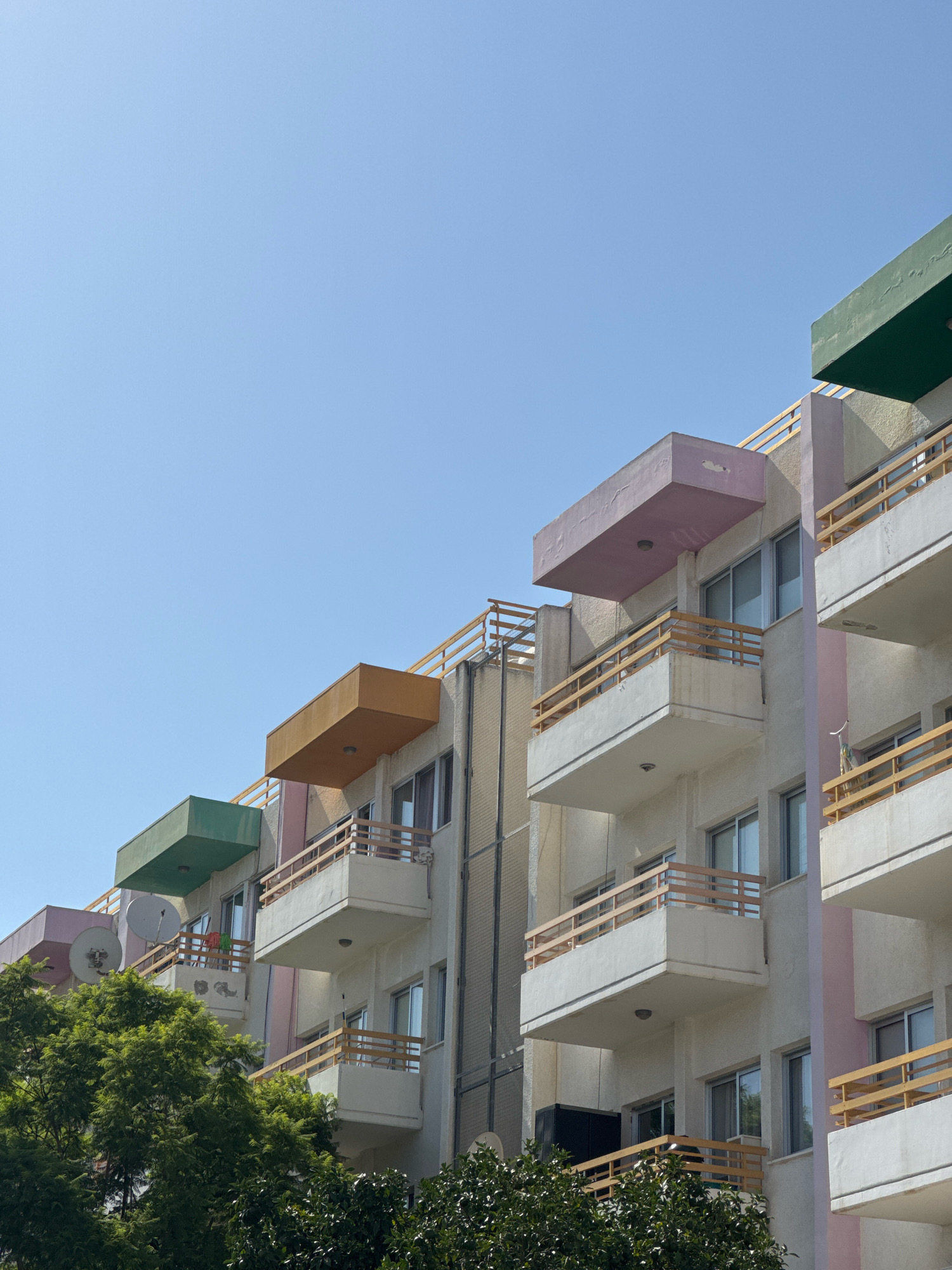 The image shows a portion of a multi-story apartment building in Germasogeia Municipality, Cyprus, against a clear blue sky. The building features multiple balconies with light-colored railings and concrete overhangs. Each overhang is painted a different pastel color: green, pink, and orange are visible. The building's facade is primarily light-colored with some pink accents. Trees with dark green foliage are visible in the lower left and bottom of the frame. Satellite dishes are mounted on some of the balconies. The architecture suggests a modern residential building.