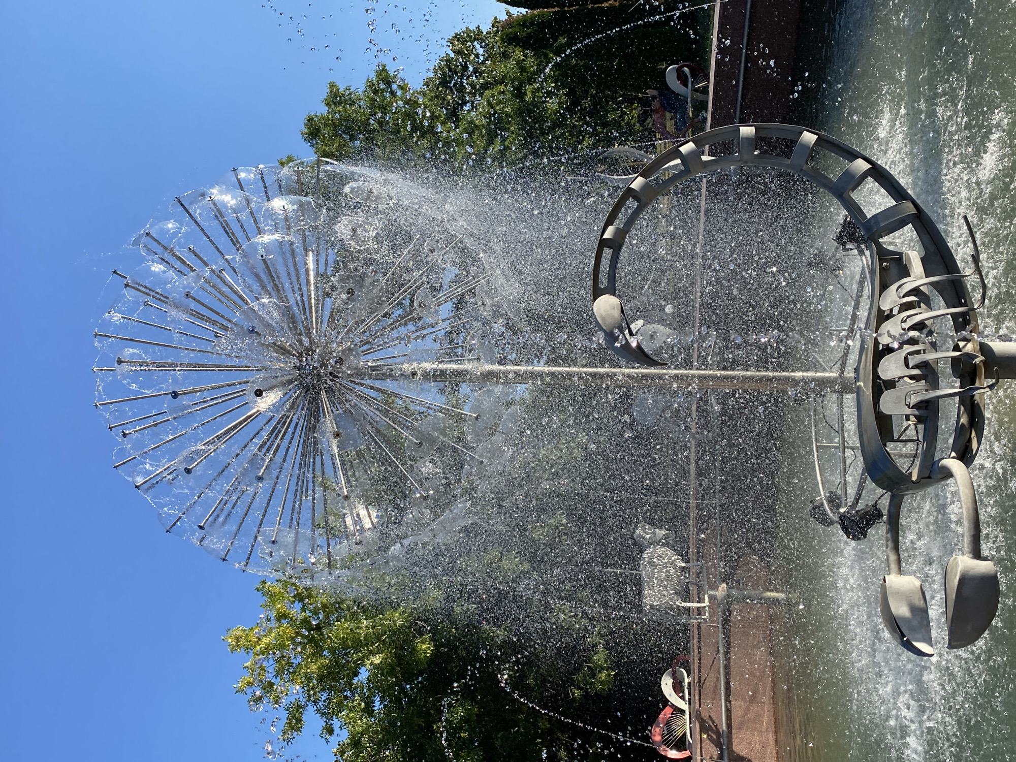The image shows a large, spherical metal sculpture with numerous radiating rods, resembling a dandelion seed head, at the center of a fountain. Water sprays outwards from the sculpture, creating a shimmering curtain of droplets. At the base of the fountain sculpture, there is a metal structure shaped like a scorpion with a circular segmented tail. In the background, there are trees and a clear blue sky. The fountain is located in the Public Garden (Grădina Publică) in Chișinău, Moldova, and the surrounding area includes sculpted animal figures, such as a metallic sheep and decorative red and white elements.