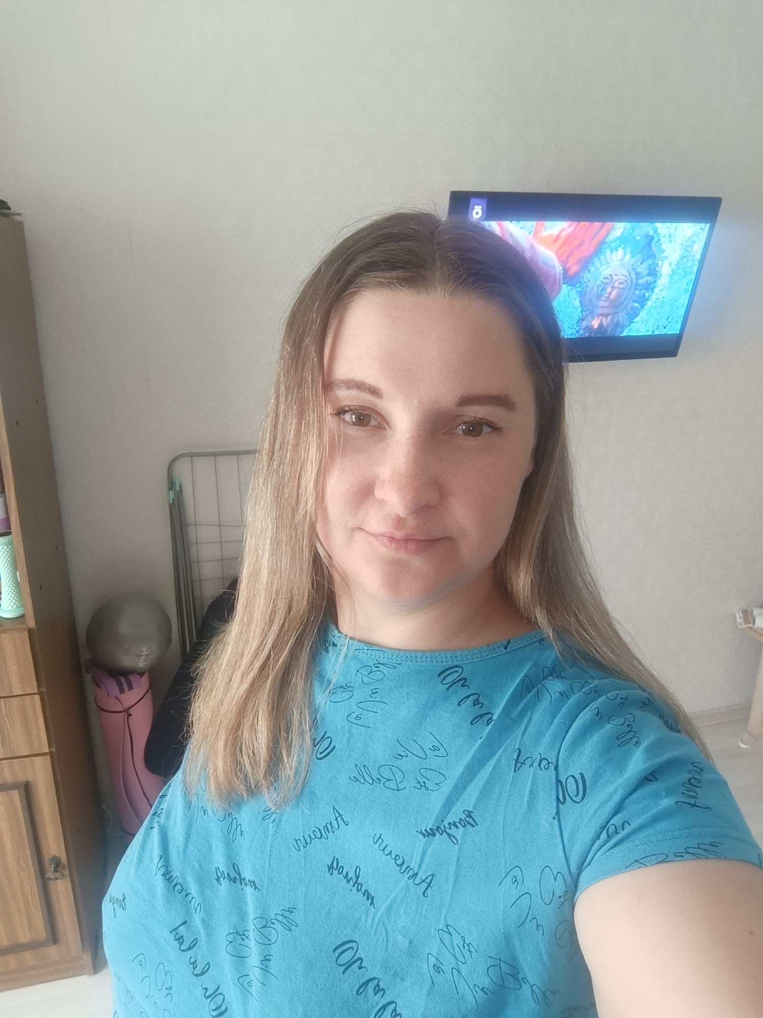 A woman is posing for a selfie indoors. She has light brown hair and is wearing a blue T-shirt with written patterns. Behind her is a wall-mounted television screen displaying an image. To the left, there is a wooden cabinet and a metal clothes drying rack. Additionally, a grey exercise ball and a pink yoga mat are visible in the background.