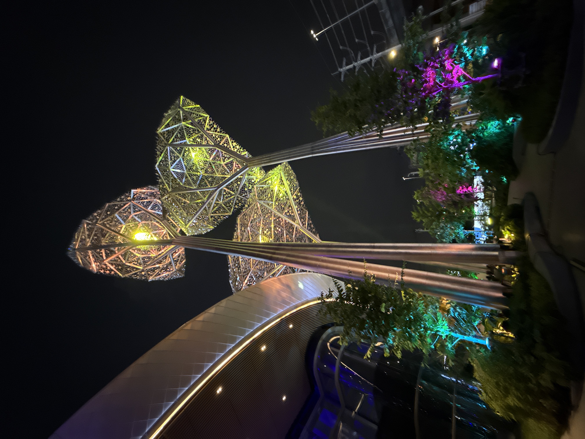 An outdoor nighttime scene features stylized metallic tree sculptures illuminated by colored lights. The sculptures have intricate, geometric canopies and tall, slender trunks. In the background, a curved, modern building with a metallic facade and integrated lighting is visible. Lush green foliage, also lit with green, blue, and purple lights, surrounds the base of the sculptures and creates a landscaped garden area. A pathway winds through the garden. The overall impression is one of a futuristic and artistically lit urban environment. The location is Bang Kaeo Town Municipality, Thailand.