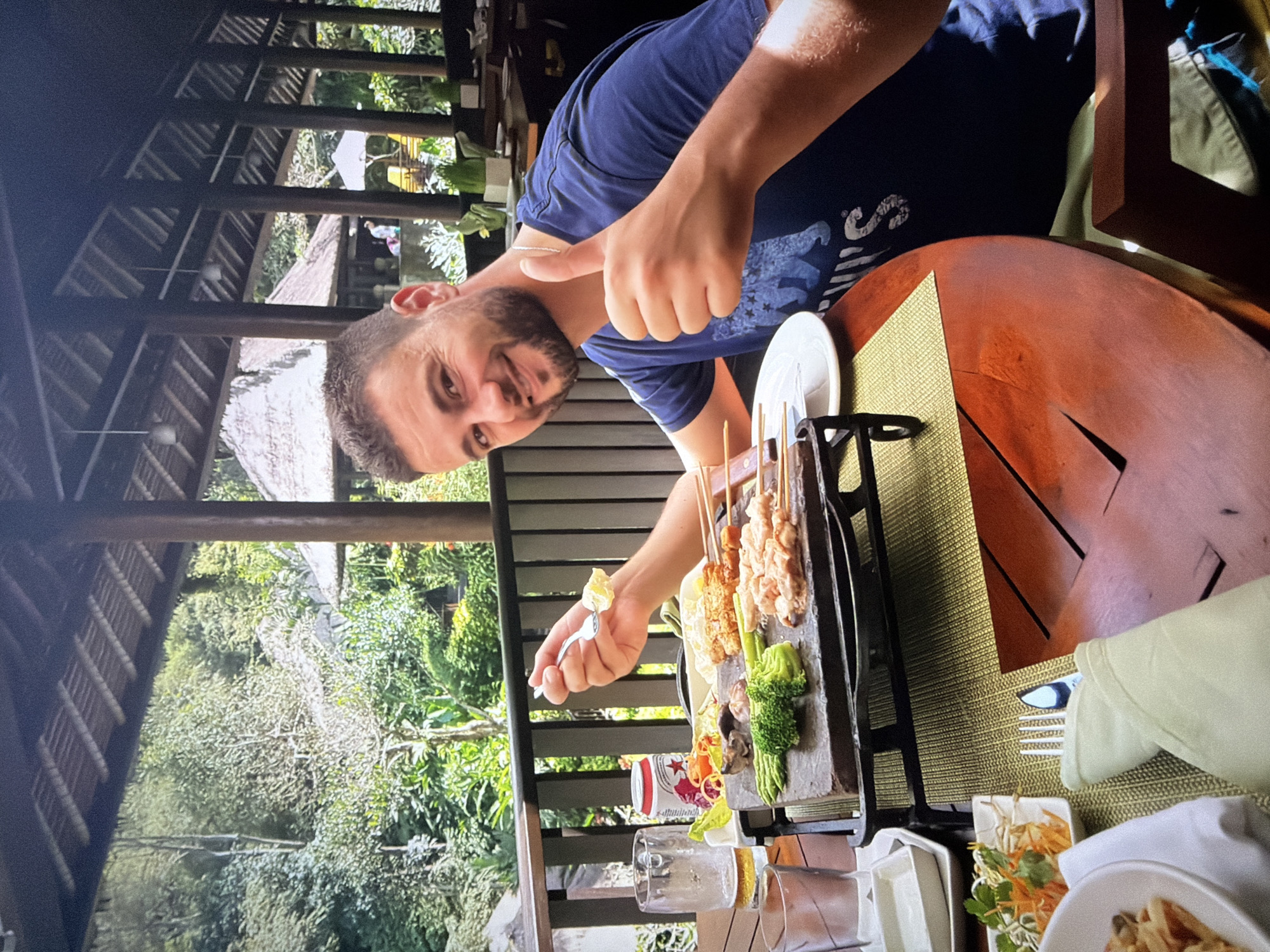 An overhead-angled shot captures a man dining outdoors in what appears to be a tropical setting. The man, with a beard and dark blue t-shirt, is positioned at the right of the frame, smiling and giving a thumbs-up gesture towards the camera. He holds a fork with a piece of food on it.
In front of him on a round wooden table, a main dish of various skewers (appearing to be chicken, shrimp, and vegetables like broccoli and asparagus) is presented on a multi-tiered stone platter, held by a black metal stand. Alongside this, there are smaller white bowls containing side dishes, such as salads or noodles, and a white plate. A woven green placemat sits beneath the food. Glasses and a partially visible red and white can are also on the table.
The background reveals a lush, green landscape dominated by trees and foliage, suggesting a vibrant natural environment. The dining area is an open-air structure, characterized by dark wooden beams and pillars, and a visible traditional-style roof in the distance. The bright natural light indicates it is daytime. The man's smile and thumbs-up convey a sense of enjoyment and satisfaction. While a location context of Pétange, Luxembourg was provided, the visual elements of the image, particularly the vegetation and building style, strongly suggest a tropical or Southeast Asian environment. No clearly decipherable text is visible on the man's shirt or the can.