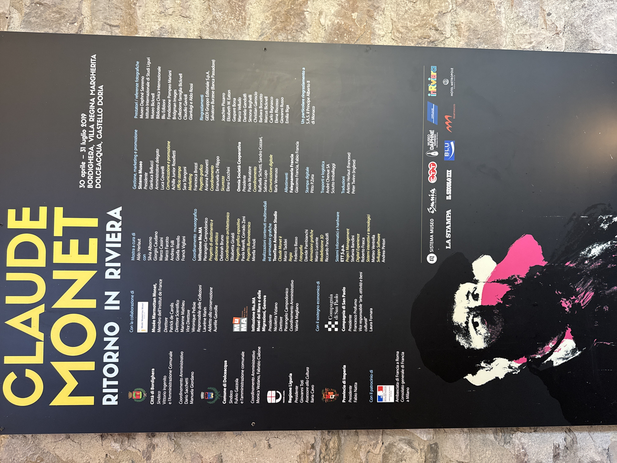 This is a photograph of a large black poster advertising an exhibition of Claude Monet's work titled "Ritorno in Riviera." The exhibition is taking place from April 30 to July 31, 2019, at various locations including Bordighera, Villa Regina Margherita, and Dolceacqua, Castello Doria. The poster features a stylized, graphic representation of Claude Monet's face in black, white, and pink. The text on the poster is primarily in white and yellow, with some smaller sections in other colors. The poster is mounted outdoors, as evidenced by the rough, textured stone wall visible to the right and behind it. The lighting suggests it is daytime. The poster itself is the main subject, with the surrounding environment providing context.