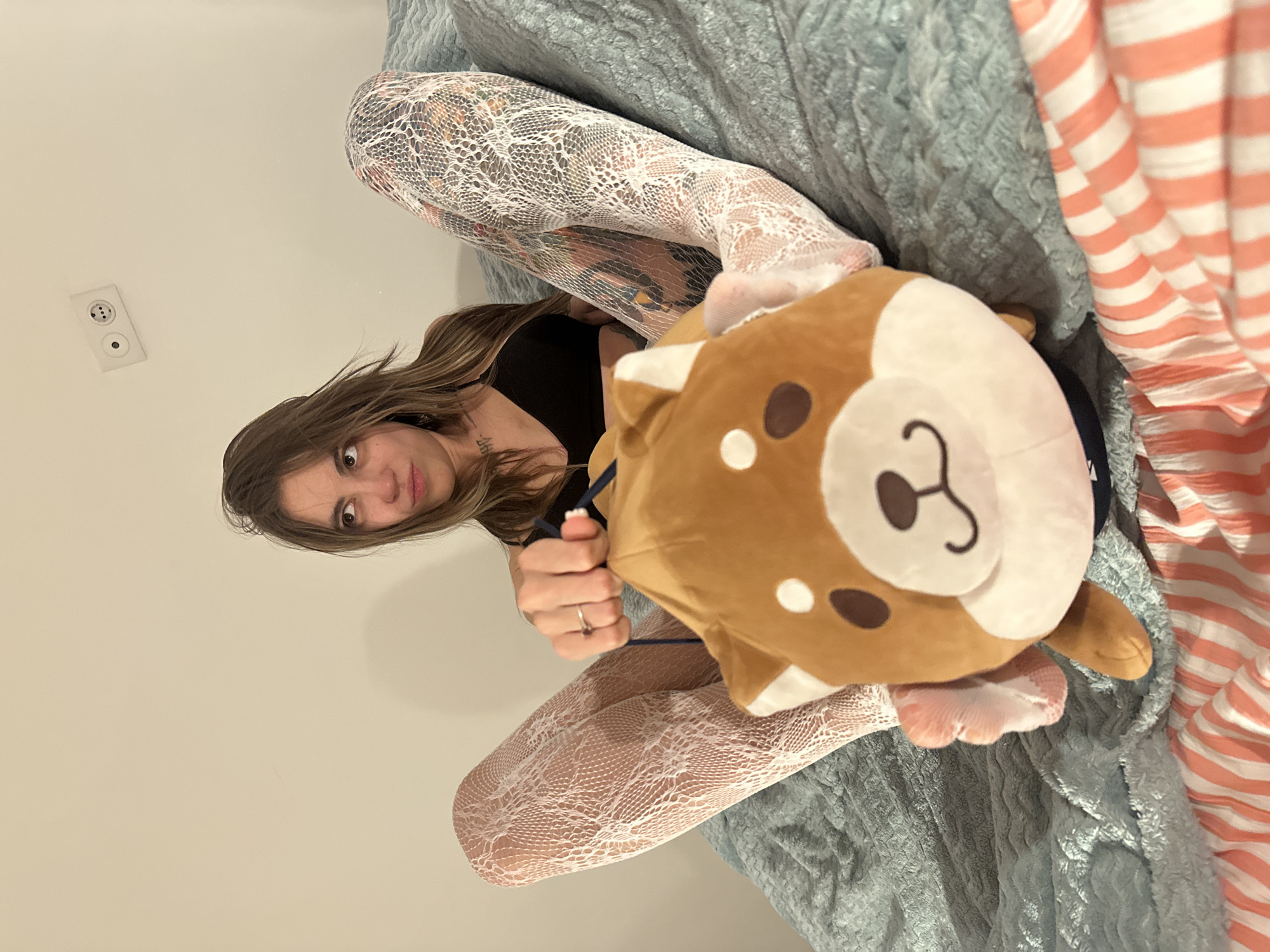 The image shows a young woman sitting on a bed, holding a stuffed animal. She is wearing a black top and white lace tights. The bed is covered with a blue textured blanket and a striped comforter. In the background, a white wall with an electrical outlet is visible. The scene appears to be indoors, likely in a bedroom. The image does not contain any specific information that would allow for the identification of its location in Moscow, Russia.