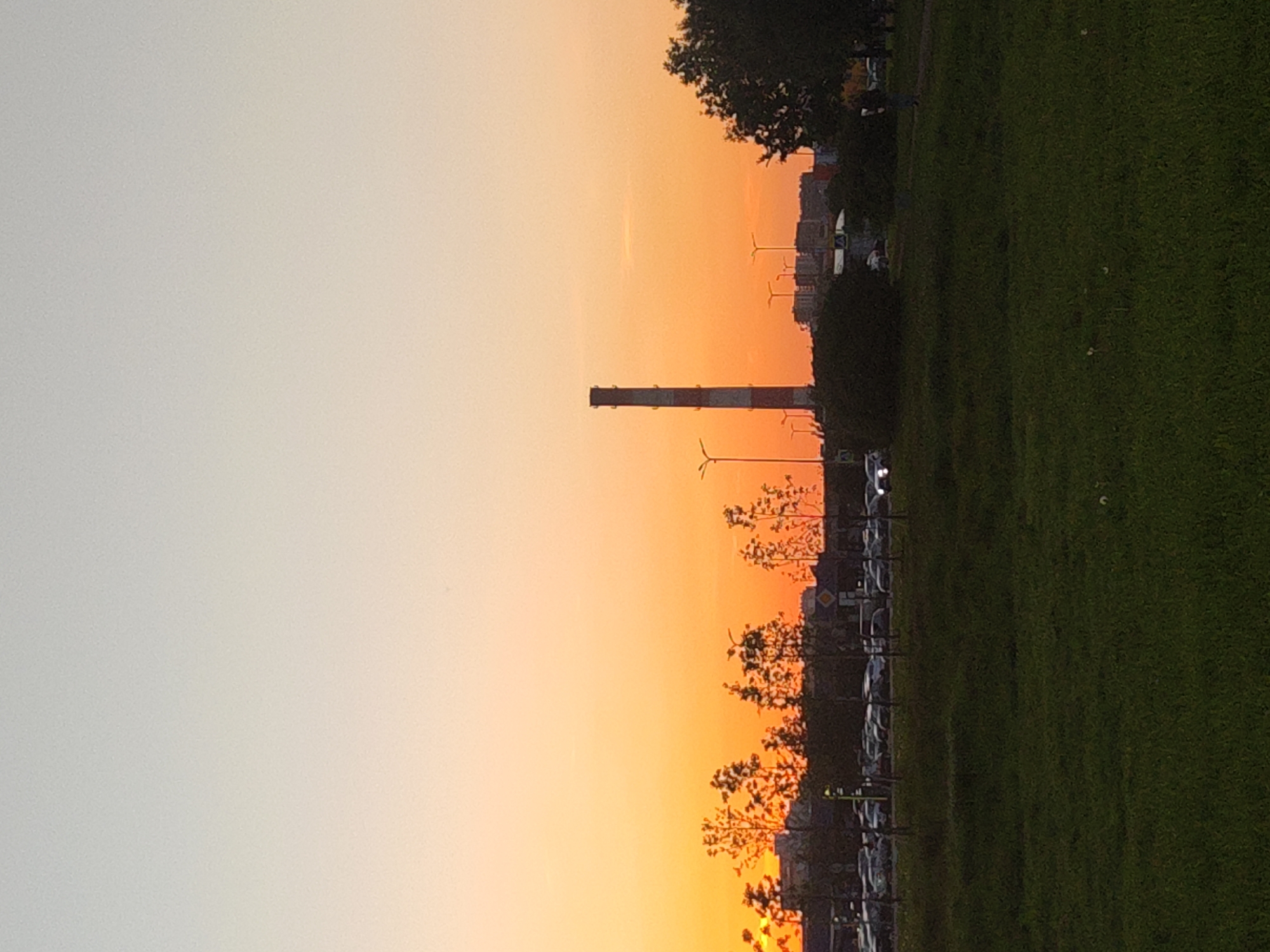 An image captured in portrait orientation (when viewed as provided, or landscape when rotated correctly) depicts an urban or park setting during sunset in Saint Petersburg, Russia.
The sky, dominating the upper portion of the scene (when upright), displays a striking gradient. It transitions from a pale, light grey on the left to vibrant orange and yellow hues on the right, clearly indicating either sunset or sunrise under clear weather conditions.
Along the horizon, several elements are silhouetted against the colourful sky. A tall, slender industrial chimney, marked with horizontal bands, stands prominently on the left. To its right, several small, two-bladed wind turbines are visible in the mid-distance. A line of trees with sparse foliage extends across the mid-ground, with some areas of denser tree canopy on the far right. Faint outlines of distant buildings form an urban skyline behind the trees.
The lower half to two-thirds of the image is an expansive, well-maintained green grass field, suggesting a park or large recreational area. Along the very bottom edge of this grassy expanse, a narrow strip appears to be a road or parking area, with visible streetlights and what seem to be parked vehicles or their lights.
No specific activity or event is taking place, and no distinct people are visible. The overall mood is tranquil, characterized by the serene beauty of the fading daylight. There is no visible text in the image.