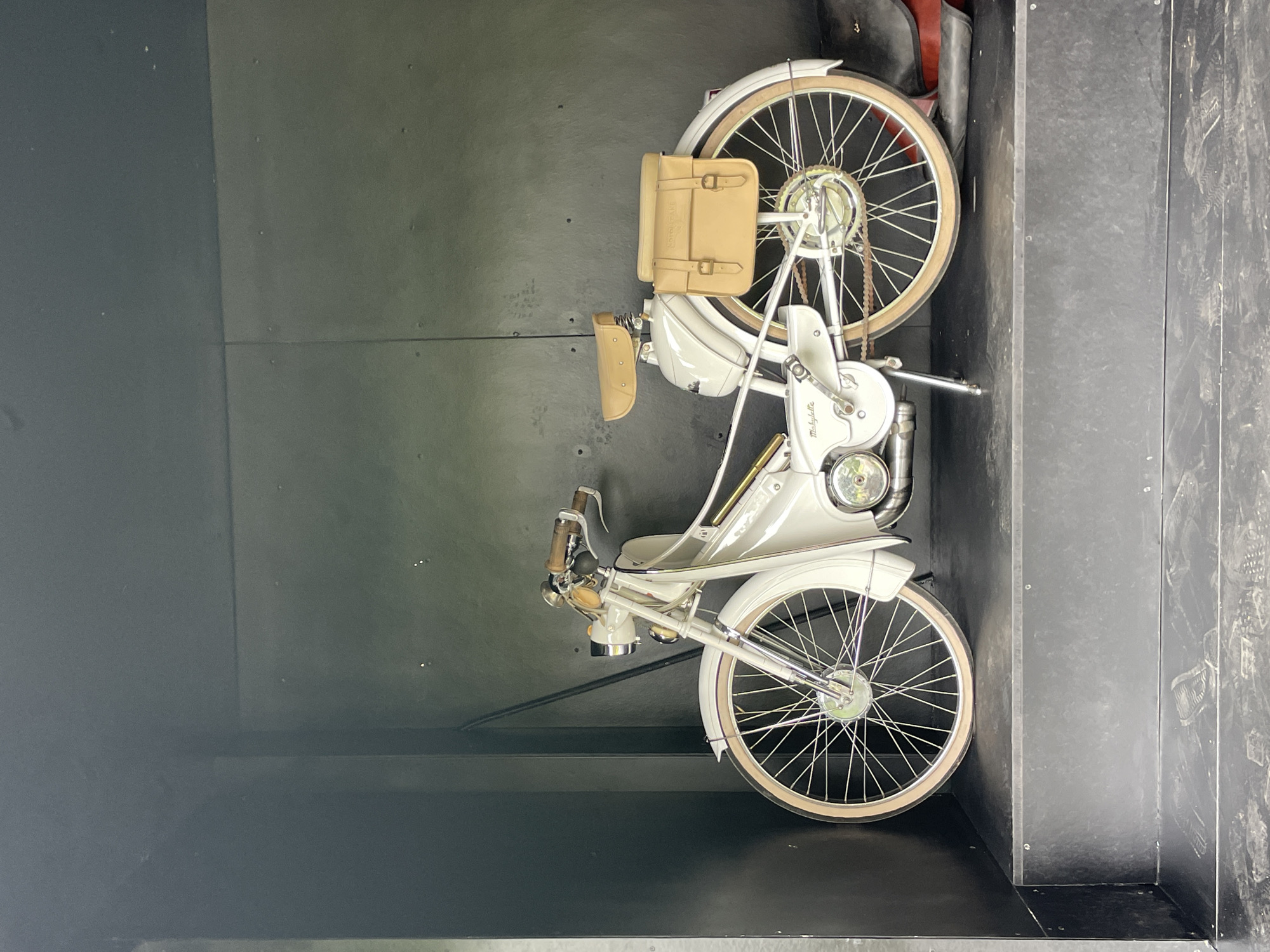 A vintage white moped is parked inside a dark, enclosed structure. The moped features tan leather saddlebags mounted on the rear fender and a tan seat. The handlebars are also tan, and the wheels have tan tires with silver spokes. The moped is on a dark platform within the structure, which has black walls and ceiling. There is no indication of the specific location within Cam Ly - Đà Lạt, Vietnam, in the image.