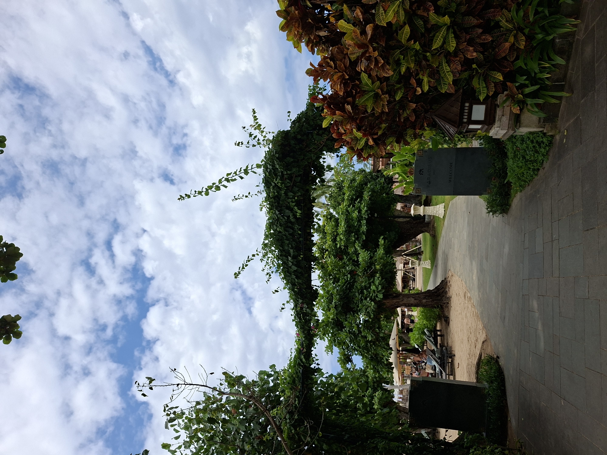 The image shows a pathway in Kuta Selatan, Indonesia, with a lush, green archway formed by trained vines and trees. The pathway is paved with gray stone tiles and lined with patches of sand. Beyond the archway, sun loungers and parasols indicate a resort or beach area. A sign reading "PARADIEUS Bali" and "WELCOME" is visible on the right. The sky above is partly cloudy with blue patches. A variety of tropical plants frame the scene. The photo is taken from a low angle, looking upwards.