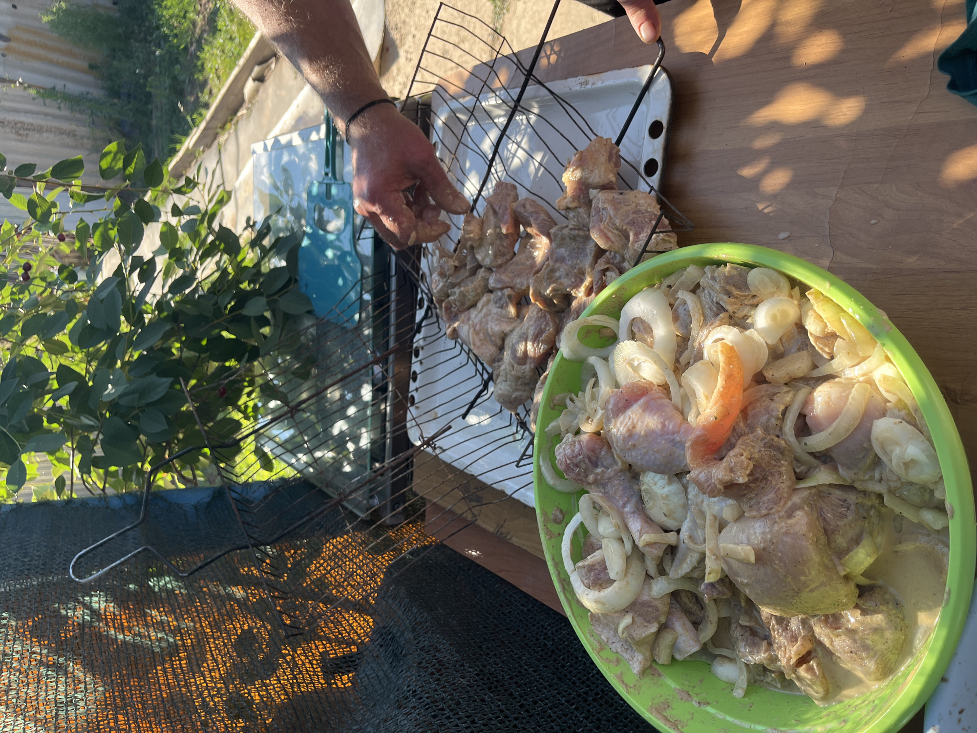 An overhead, eye-level shot captures a close-up of raw chicken pieces marinated in a creamy sauce with sliced onions, sitting in a bright green bowl. The chicken is partially cooked and arranged on a metal grill rack. A man's hand, with a black wristband, is visible, reaching to adjust the grill rack. The background reveals a sun-drenched yard with green foliage and a wooden fence, suggesting an outdoor setting. The lighting is bright and casts distinct shadows, hinting at a sunny afternoon.
