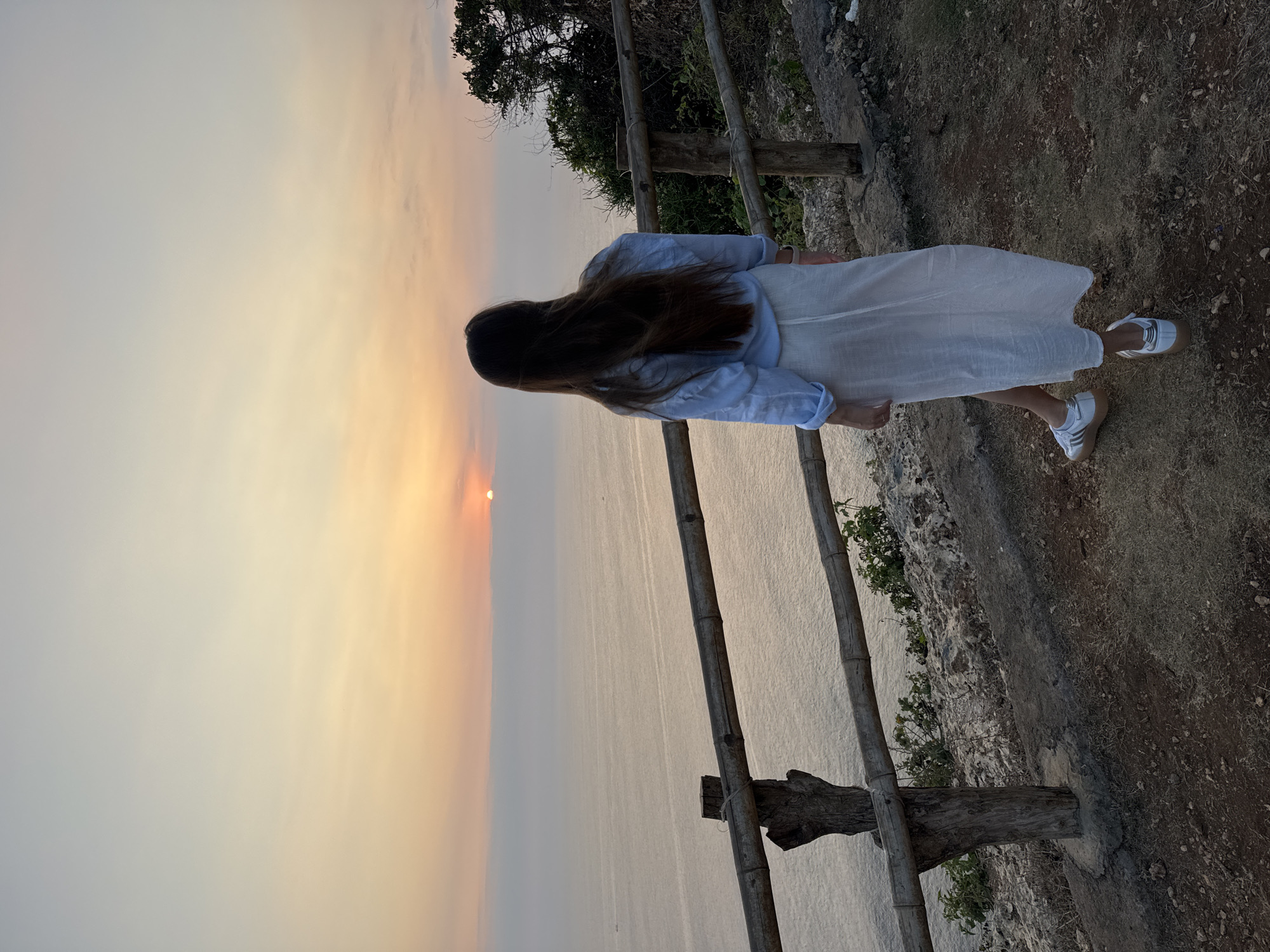 A woman stands with her back to the camera, looking out at a sunset over the ocean. She has long, dark hair and is wearing a light blue long-sleeved shirt and a white, flowing skirt. She is also wearing white sneakers. She is standing on a dirt path next to a wooden fence. The ocean is calm, and there are a few boats in the distance. The sky is filled with warm colors from the sunset, with hues of orange, pink, and yellow. The location is an outdoor vantage point overlooking a body of water at dusk.