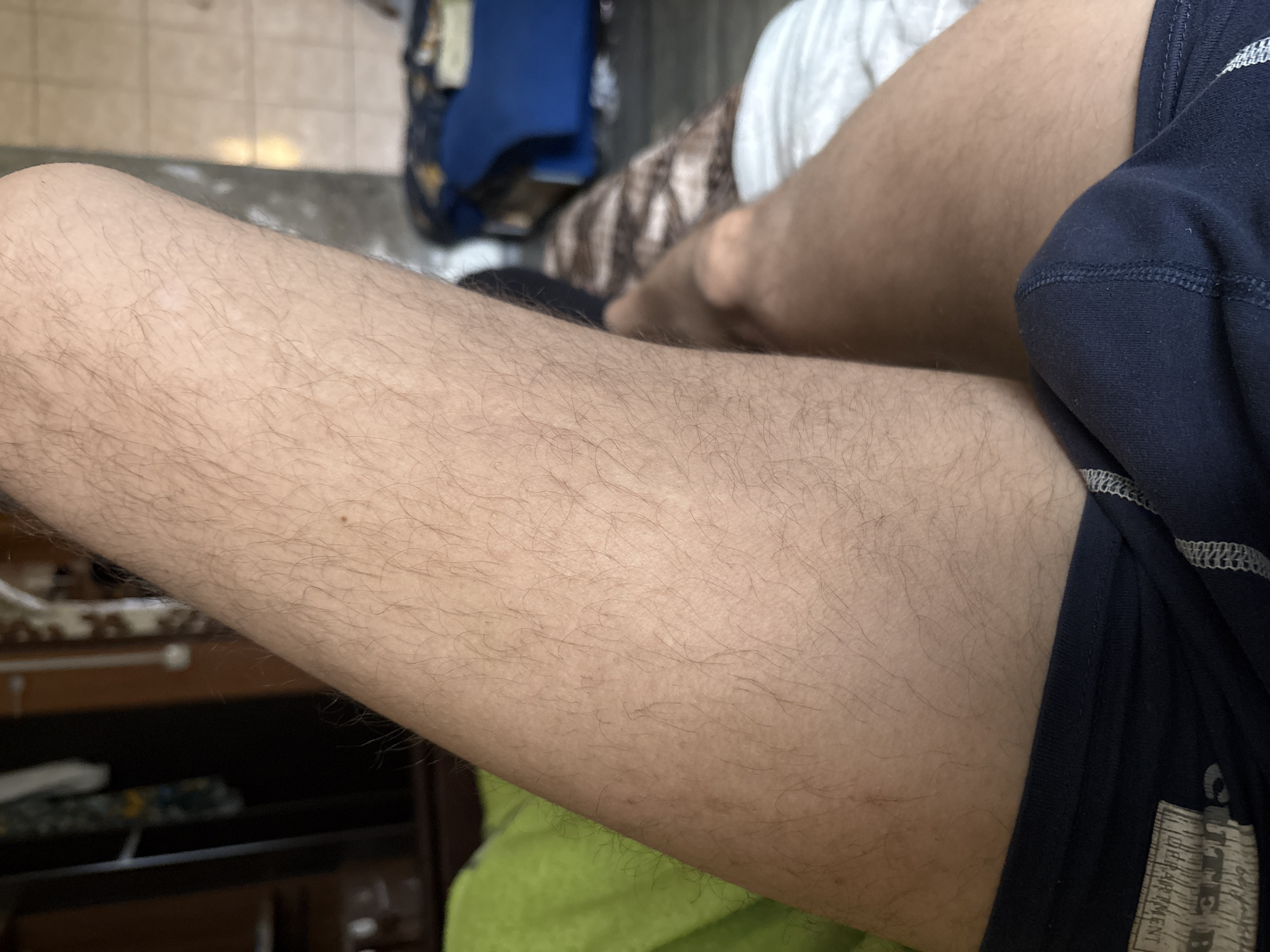 A close-up, eye-level shot focuses on a person's thigh and groin area, covered by navy blue underwear with white stitching. The thigh is covered in light brown hair. The background is blurred, showing a green blanket on the left, a bed with white sheets on the right, and a blue piece of furniture in the distance. The overall scene appears to be indoors, likely in a bedroom. There is no visible indication of the location in L'gov, Russia.