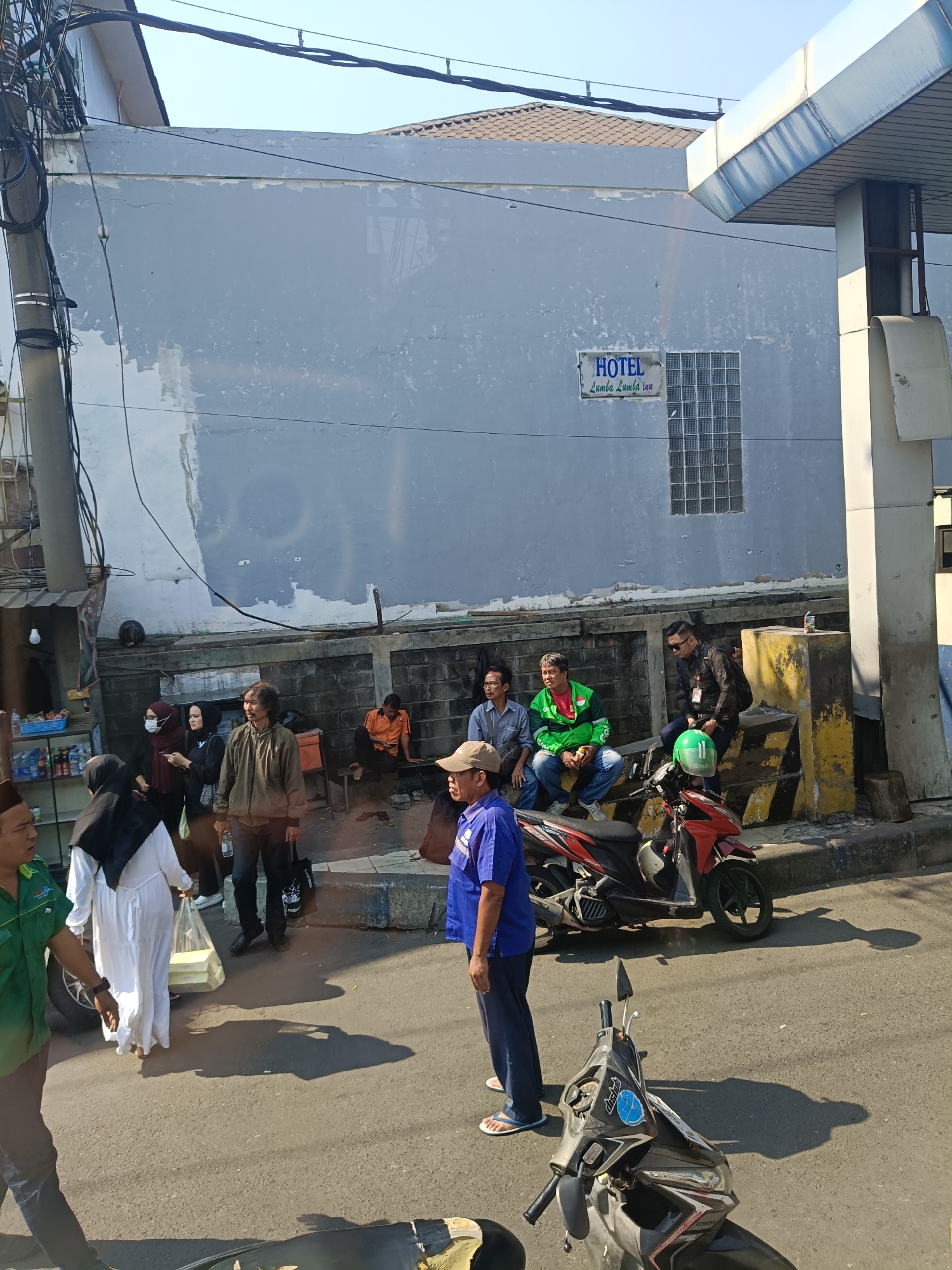 A daytime, eye-level, medium shot captures a street scene in front of a building in Kedungrejo, Indonesia. The building, painted a weathered pale blue, has a sign that reads "HOTEL Lumba Lumba Indah." To the right of the building is a white, overhanging structure with a metal roof, resembling an awning or a part of a larger building. In the foreground, a street with visible road markings stretches across the bottom of the frame. Several people are present, some sitting on a low wall in front of the building, while others are standing or walking. Two motorcycles are parked on the street. The environment appears to be a commercial or mixed-use area with a clear sky overhead.