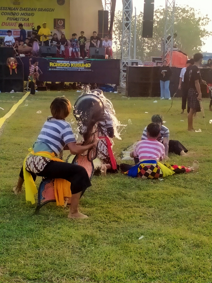 This image captures an outdoor traditional Jaranan (Kuda Lumping) performance taking place on a green grassy field in Glonggong, Sragen Regency, Indonesia. The central action in the foreground features a barefoot individual, wearing a blue and white striped shirt, black pants, and yellow and light blue fabric sashes, actively pouring water from a black bucket over the head of another performer. This second performer, adorned in traditional Javanese attire including batik fabric and a red sash, is positioned to receive the water, creating a prominent splash. Adjacent to these two, an ornate horse-shaped prop, characteristic of Jaranan, is visible. To the right, two individuals are seated on the grass; one wears a pink and white striped shirt and checkered pants, facing away from the viewer, while the other, partially obscured, has green-tinted hair and a dark shirt. In the middle ground, a yellow line or tape runs horizontally. The background features a raised stage structure with two large banners; the upper one reads "KESENIAN DAERAH SRAGEN CONDRO MOWO PINASTIKA DUWE AWE," and the lower one states "KESENIAN JARAN DOR RONGGO KUSUMO." Large audio speakers are mounted on metal truss towers flanking the stage. Various individuals are present on and around the stage, including a person in a yellow shirt on the stage and another individual in a black shirt and plaid shorts holding a long, thin implement (possibly a whip) on the grass to the right. The scene is illuminated by natural light, suggesting late afternoon.