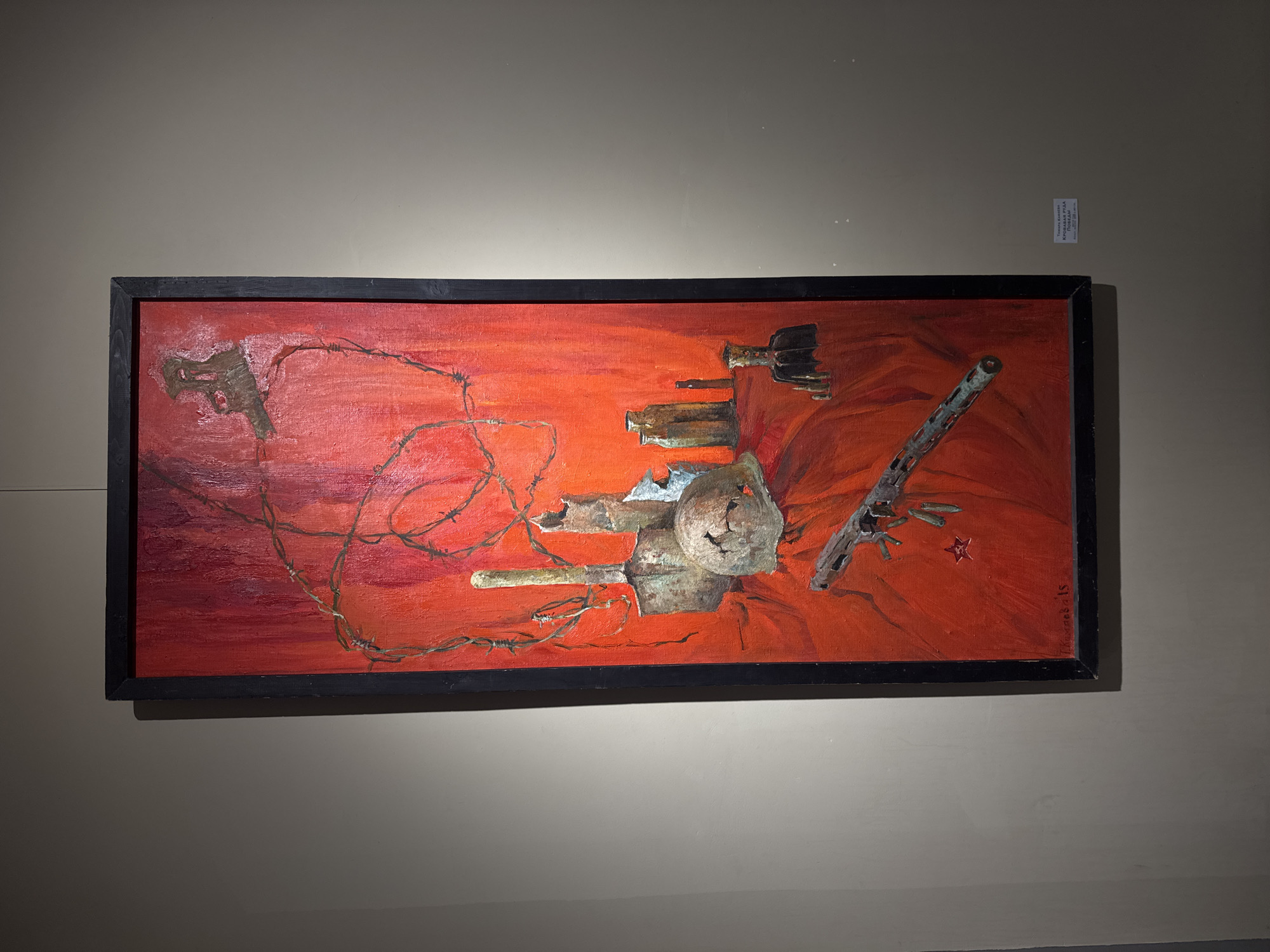 The image shows a painting hanging on a wall. The painting, framed in black, features a mostly red color palette with depictions of various objects including a gun, barbed wire, a helmet, and a rifle. These elements are rendered in a somewhat abstract style. The wall on which the painting hangs is a neutral color. A small label is visible near the upper right corner of the painting. The image appears to be taken indoors. I have no information about the location being in the city of Saint Petersburg, Russia.