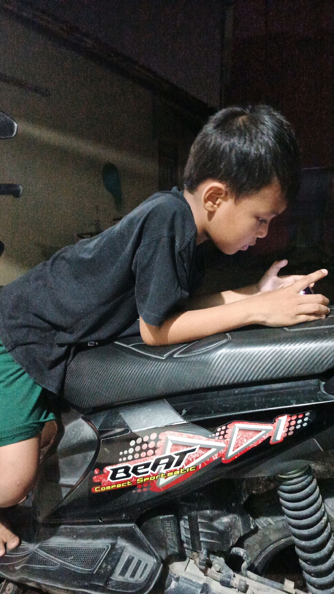 The image shows a young boy with dark hair wearing a black t-shirt and green shorts, leaning on a black motorcycle. He is looking down at a purple object in his hands, possibly a phone, and appears to be interacting with it. The motorcycle has decals on its side that read "Beat" and "Compact Sportmatic". The background is dark and appears to be indoors or a covered outdoor area, with a textured wall. The lighting is dim, with some light sources casting shadows. The scene suggests a casual, everyday moment. The image is tagged as being from Luwungragi, Indonesia.
